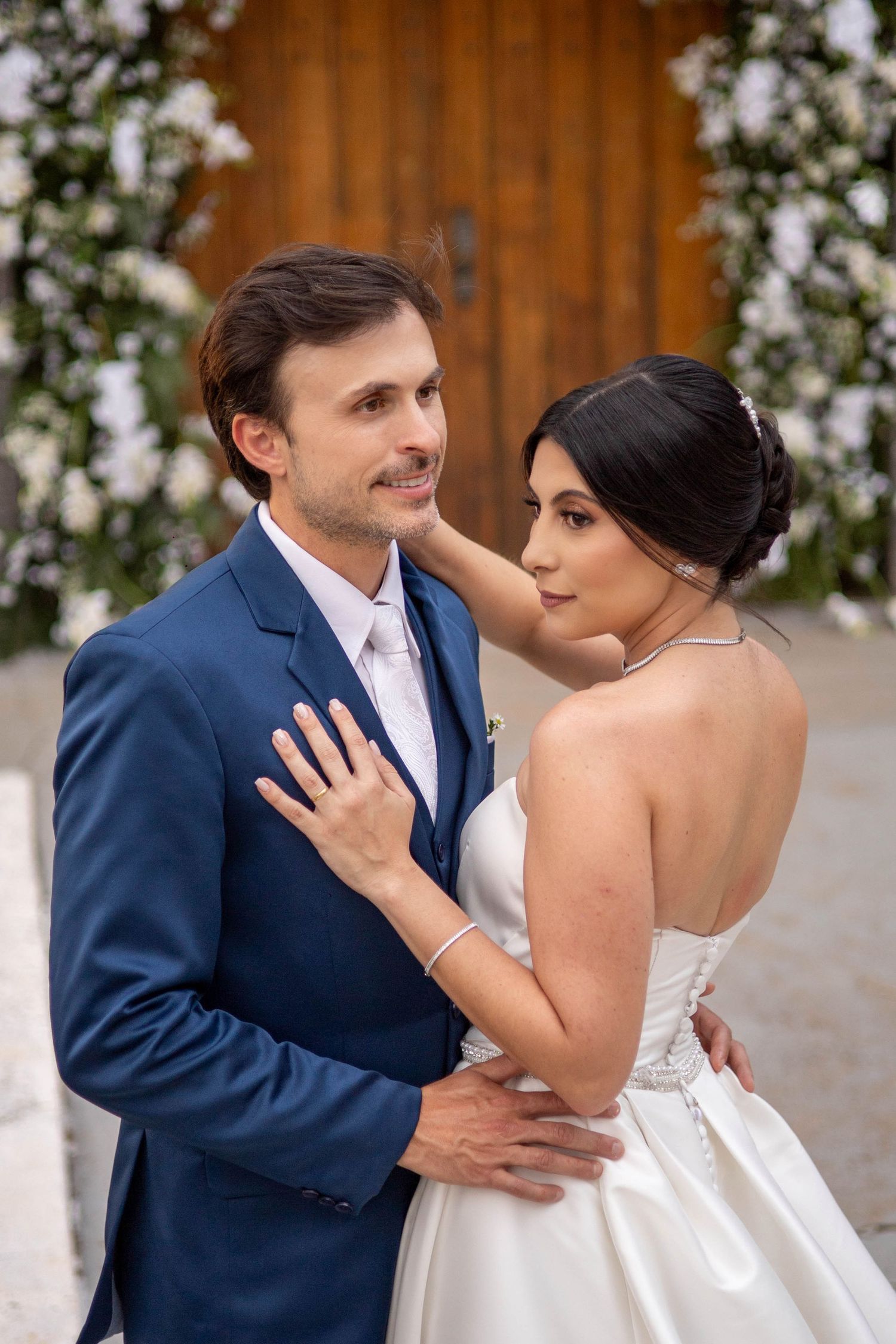A bride and groom embrace in front of a floral arch and wooden door, wearing a white gown and blue suit.