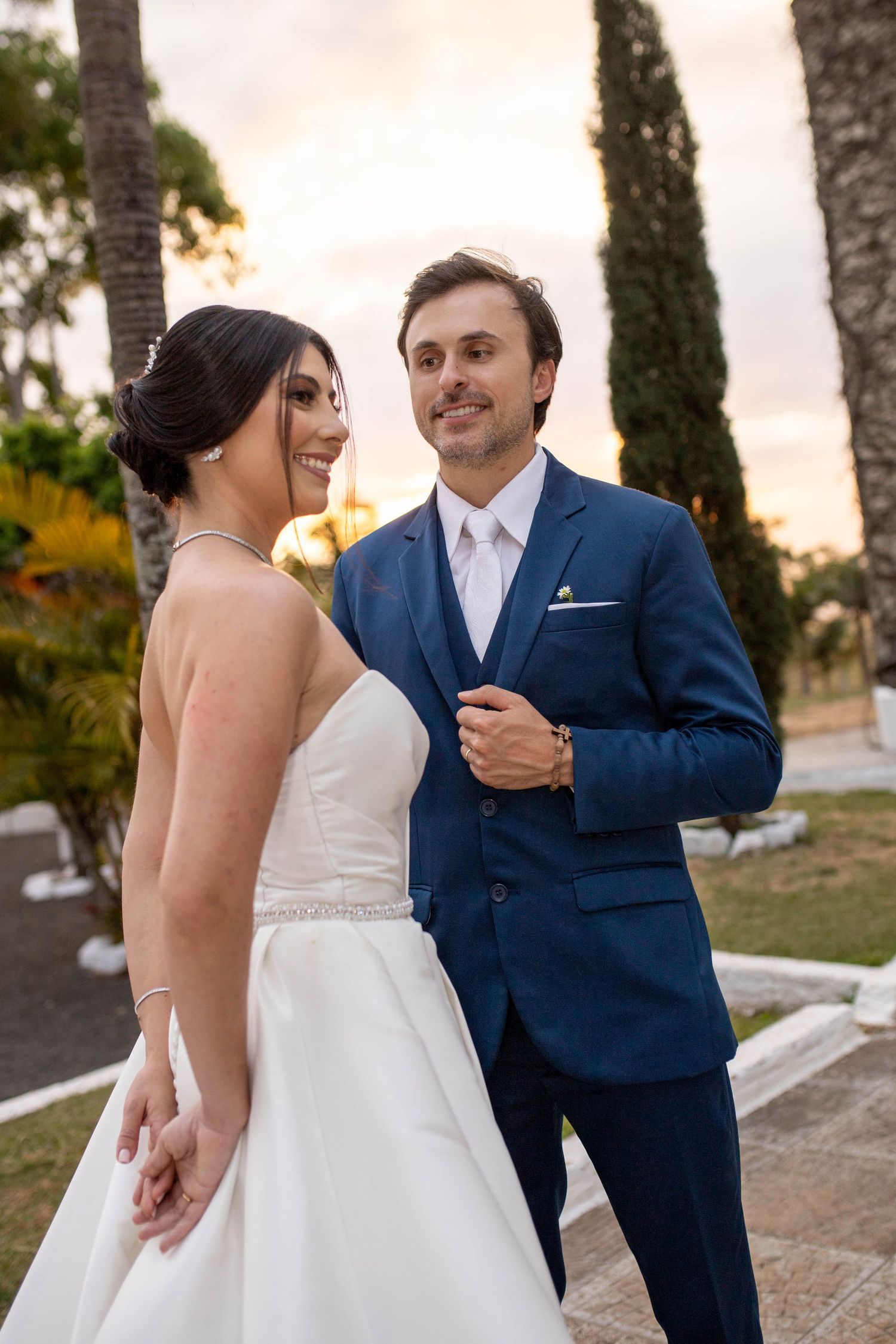 Bride in white gown and groom in blue suit share a romantic moment outdoors at sunset with palm trees.
