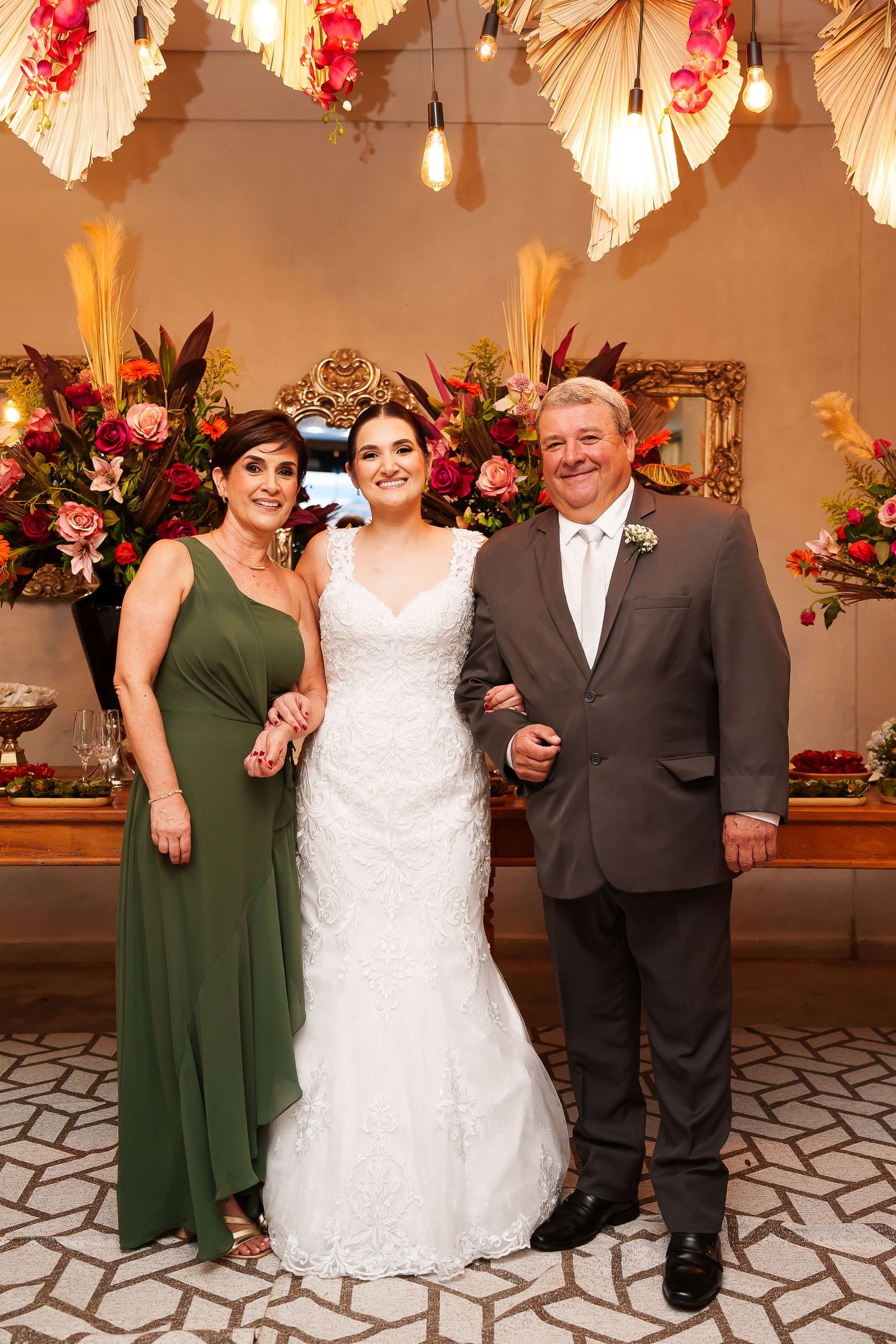 Bride in white gown poses with parents in green dress and gray suit at elegant floral wedding reception.