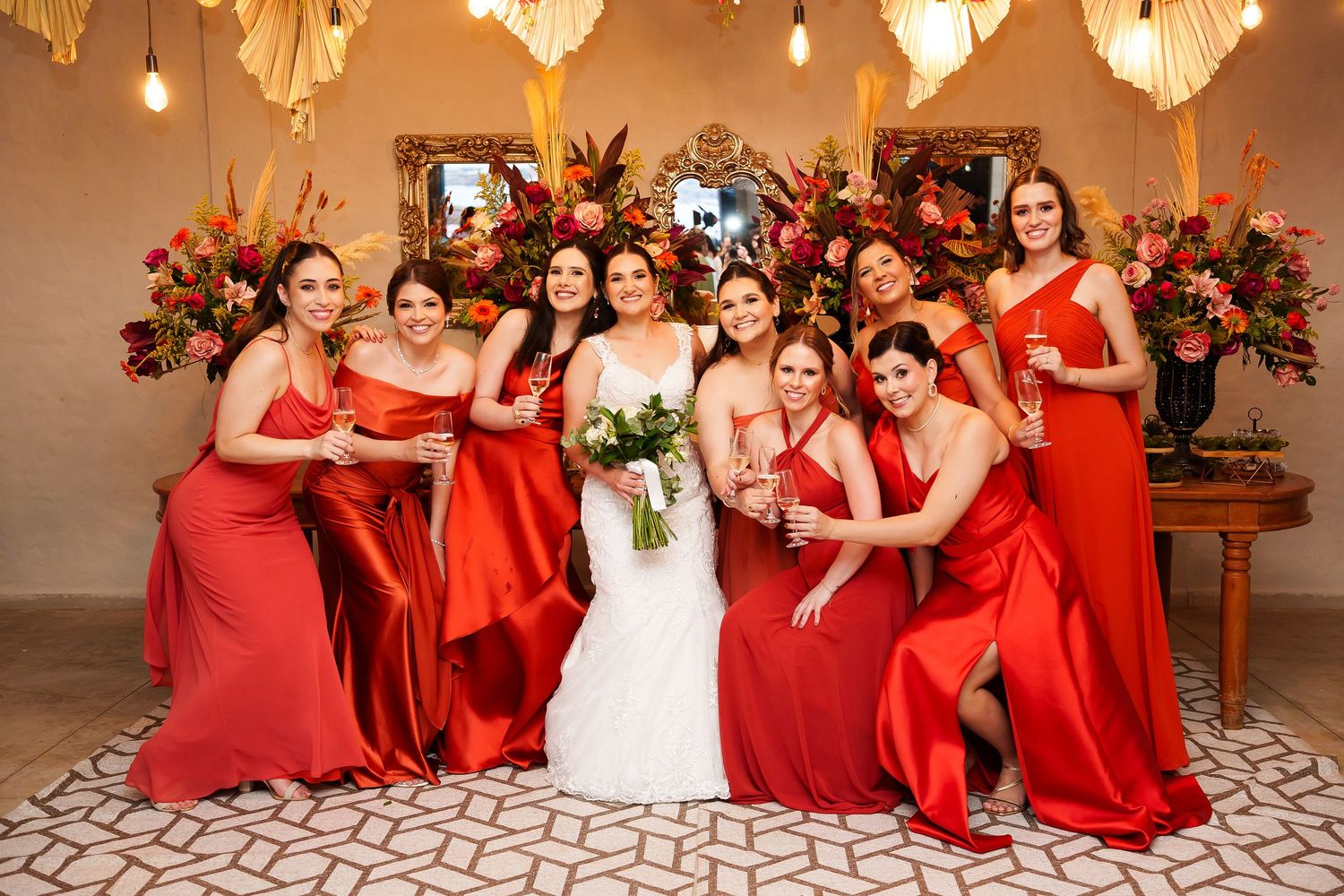 Bride in white gown poses with seven bridesmaids in red satin dresses, holding bouquets and champagne glasses indoors.