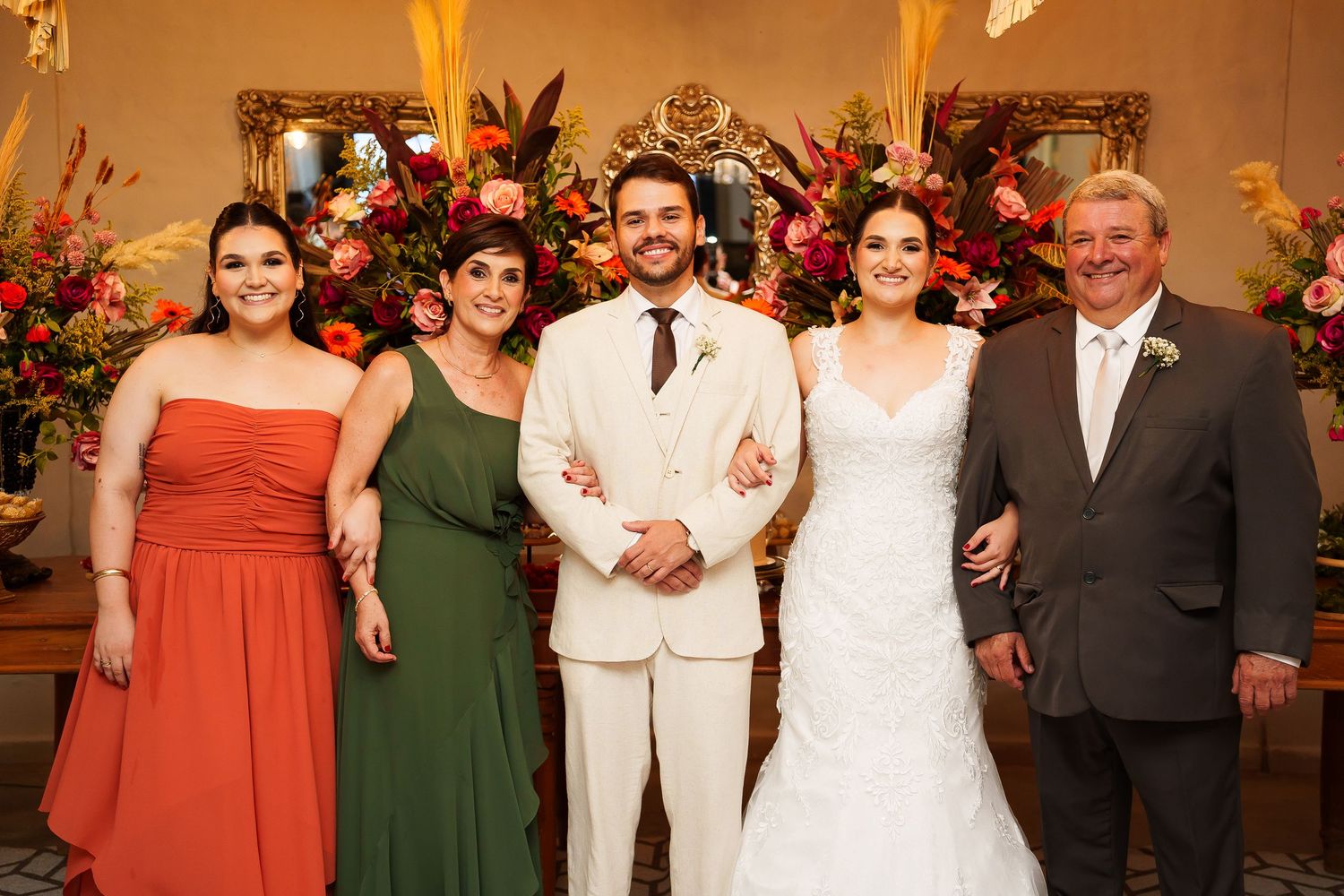 Wedding party of five posing in front of floral arrangements, bride in white gown, groom in cream suit, guests in formal attire.