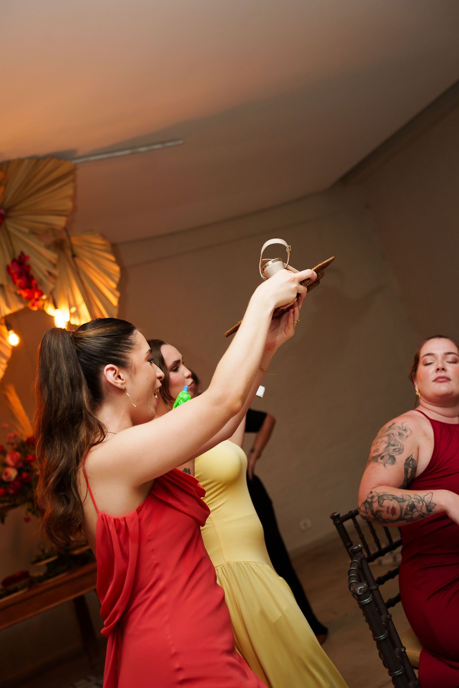 Women in red and yellow dresses celebrate at a wedding reception, one raising a champagne glass in a festive toast.