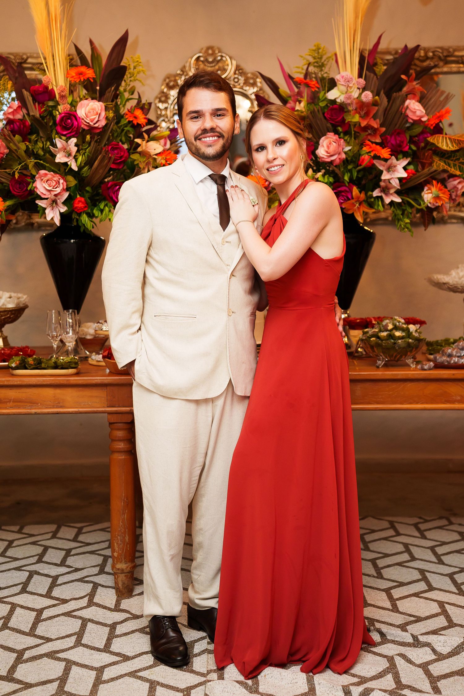 Elegant couple posing at a formal event, man in cream suit, woman in red gown, floral arrangements behind them.