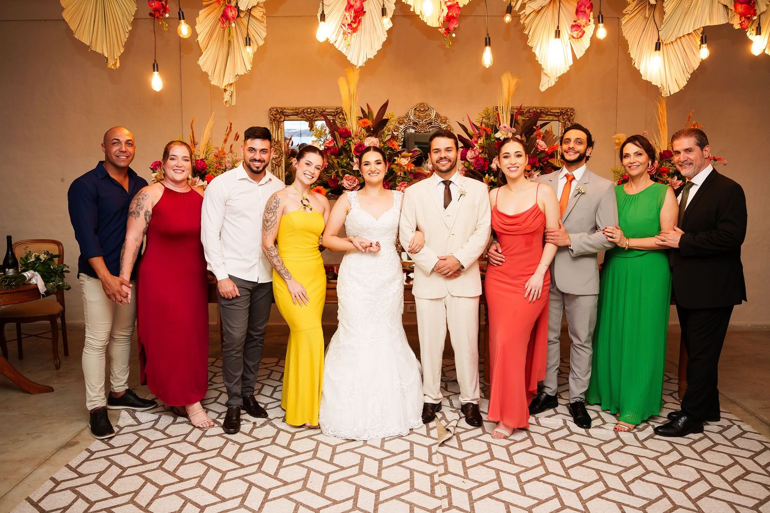 Wedding party group photo with bride in white gown, groom in gray suit, and guests in colorful formal attire indoors.