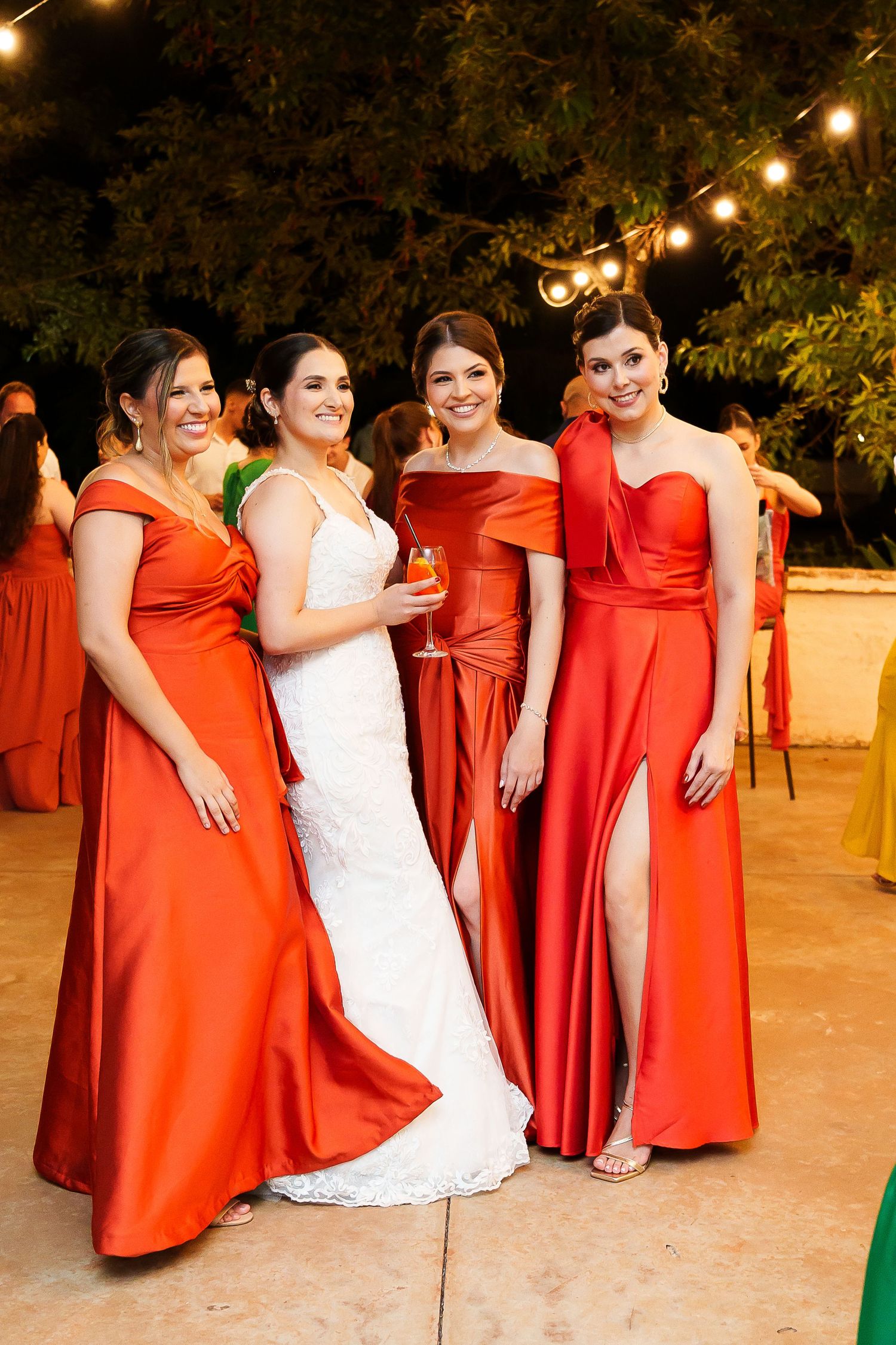 Bride in white gown poses with three bridesmaids in red satin dresses at outdoor evening wedding reception.