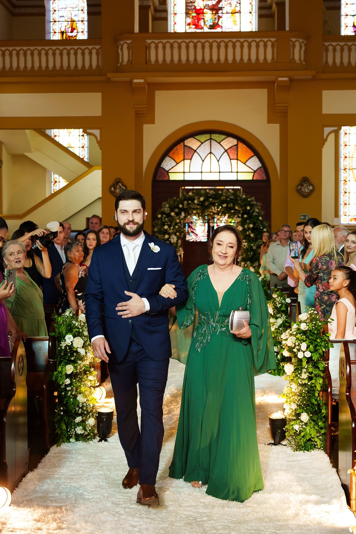 Groom in navy suit walks church aisle with woman in green gown during wedding ceremony.