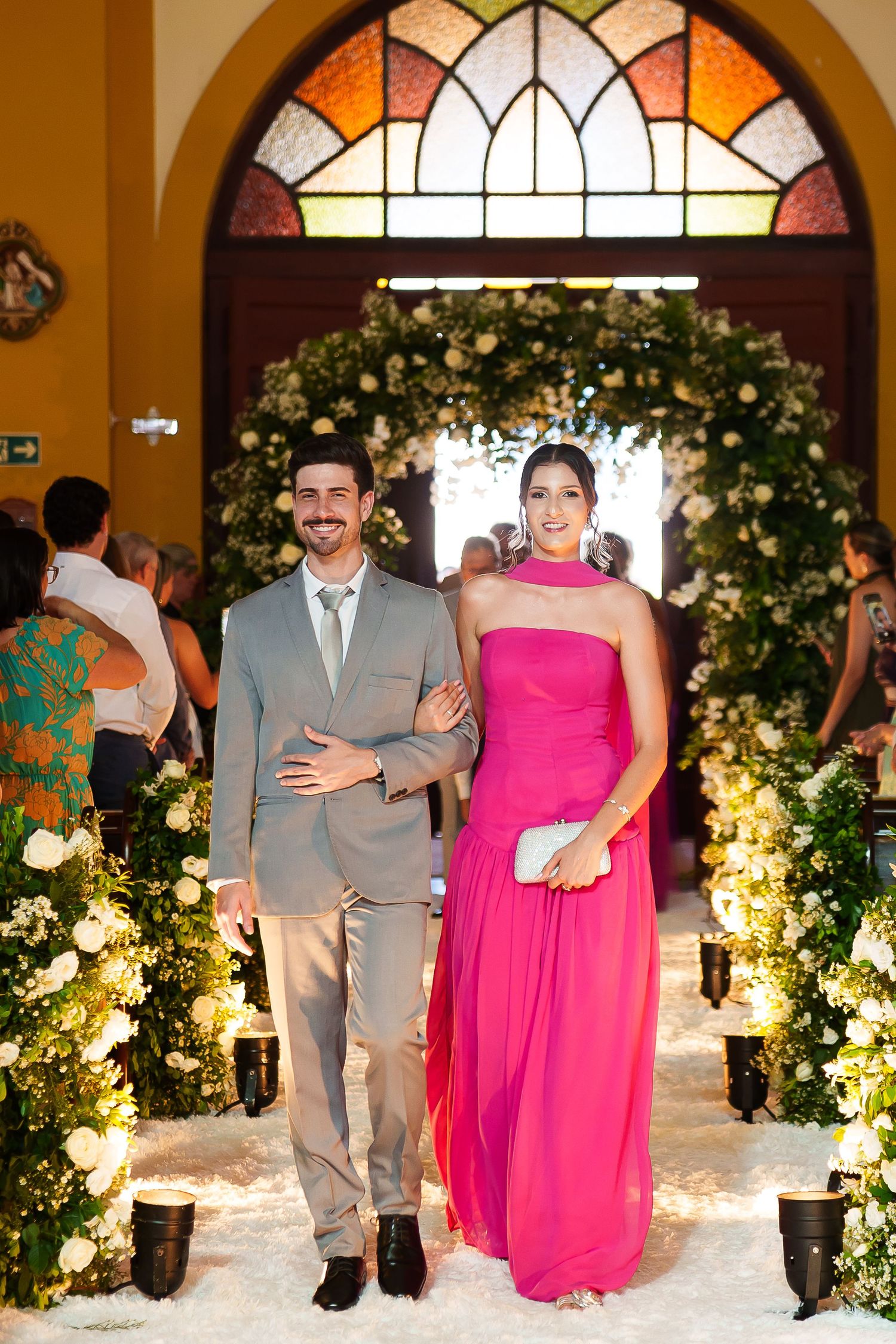 Elegant couple posing at a wedding, man in gray suit, woman in pink strapless gown, floral arch backdrop.