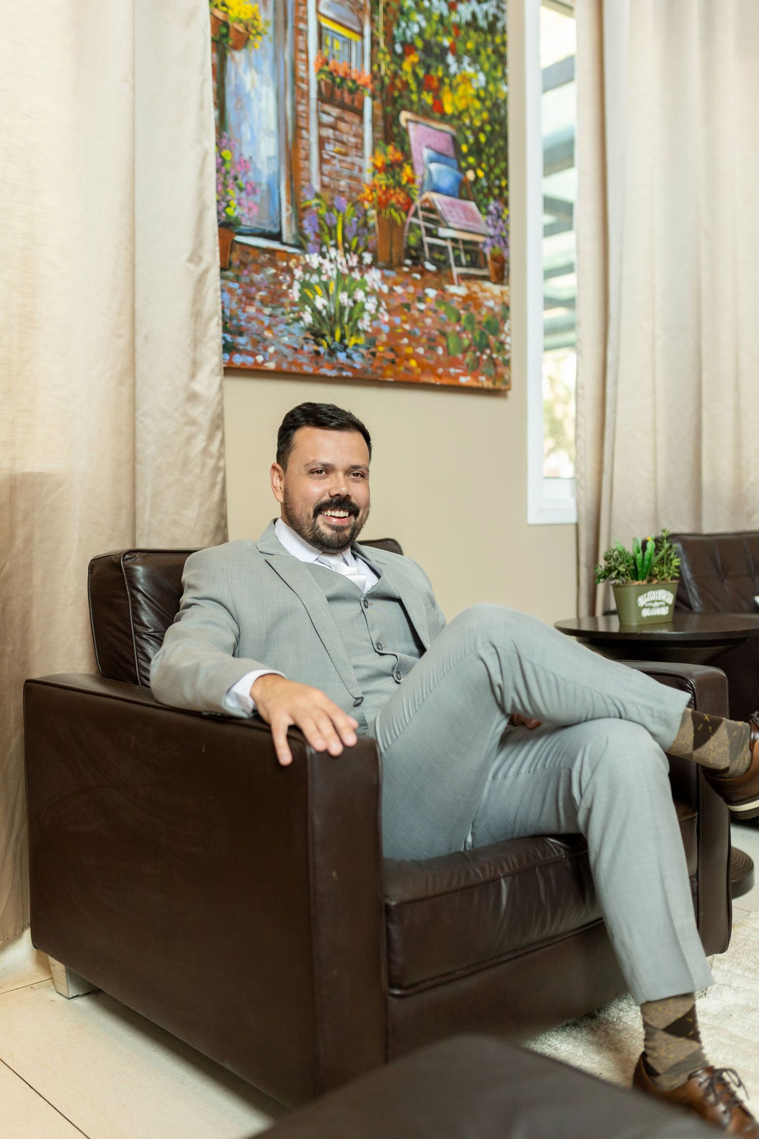 Confident businessman in gray suit relaxing in brown leather armchair beside colorful floral painting.