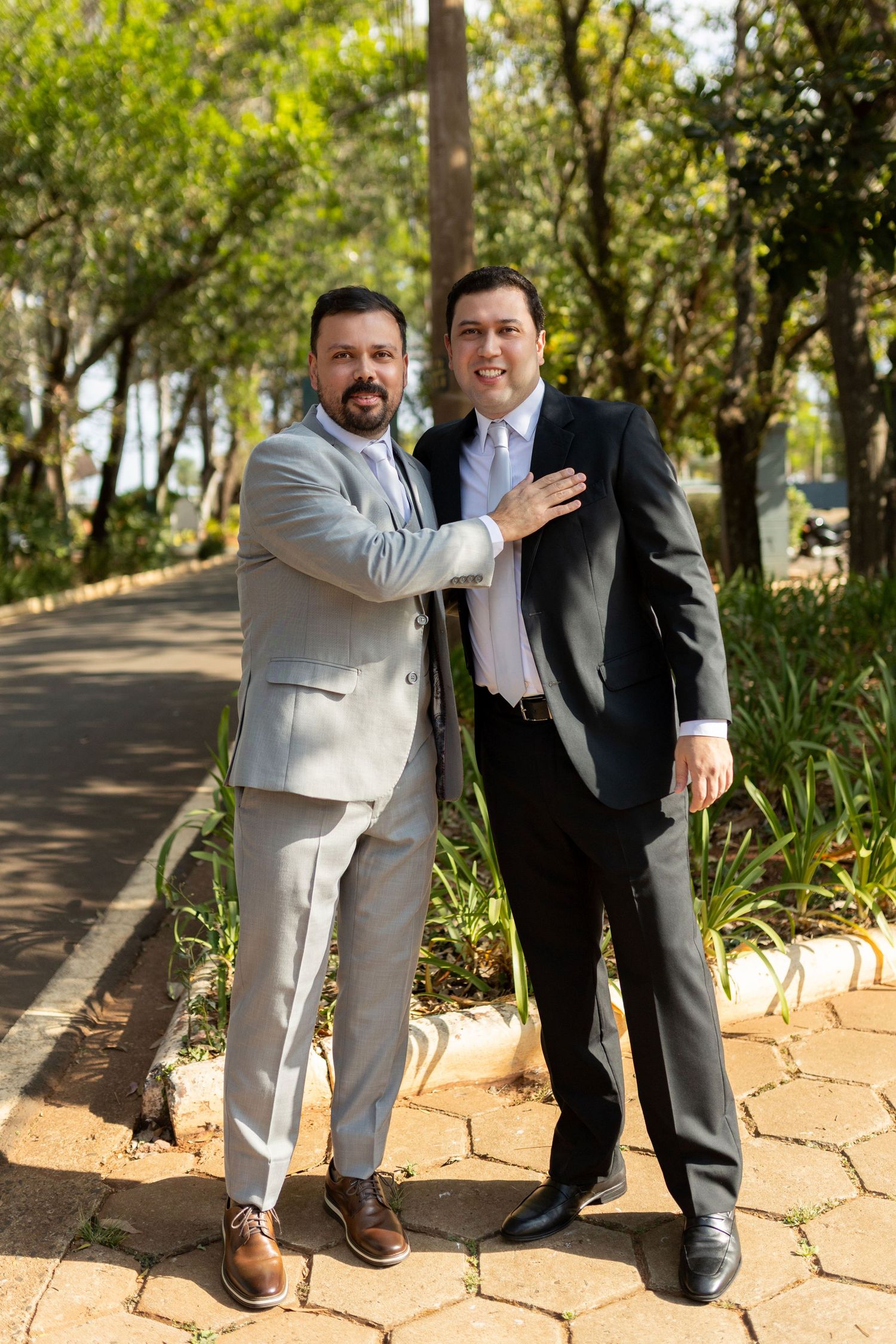 Two well-dressed men in suits posing together outdoors in a tree-lined park pathway on a sunny day.