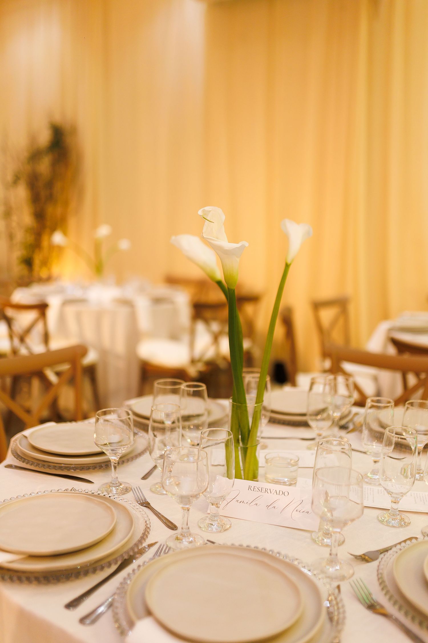 Elegant wedding reception table with white calla lilies centerpiece, crystal glasses, and gold-rimmed plates in warm lighting.