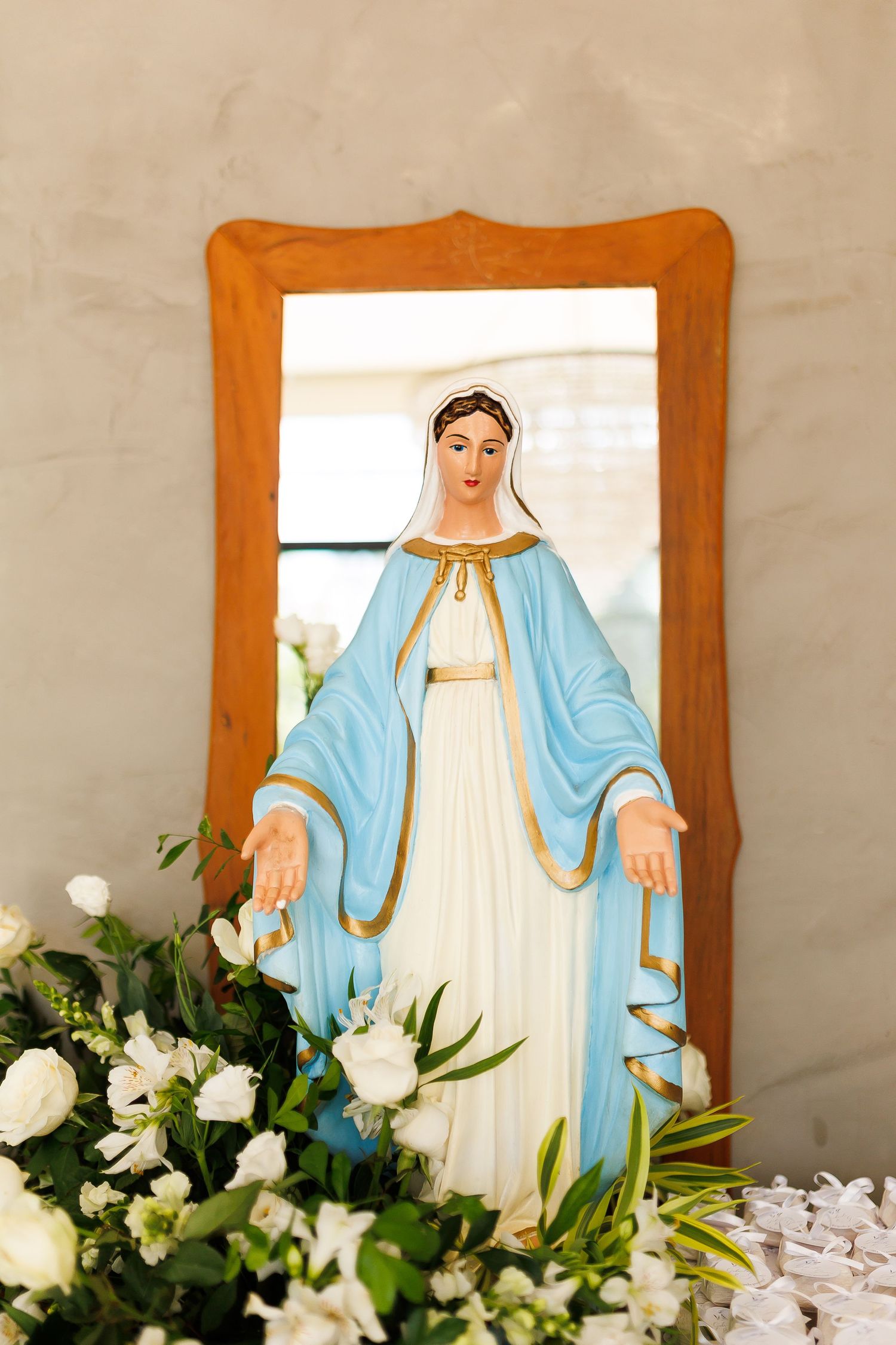 Statue of Virgin Mary in blue and white robes with gold trim, framed in wood, surrounded by white flowers.