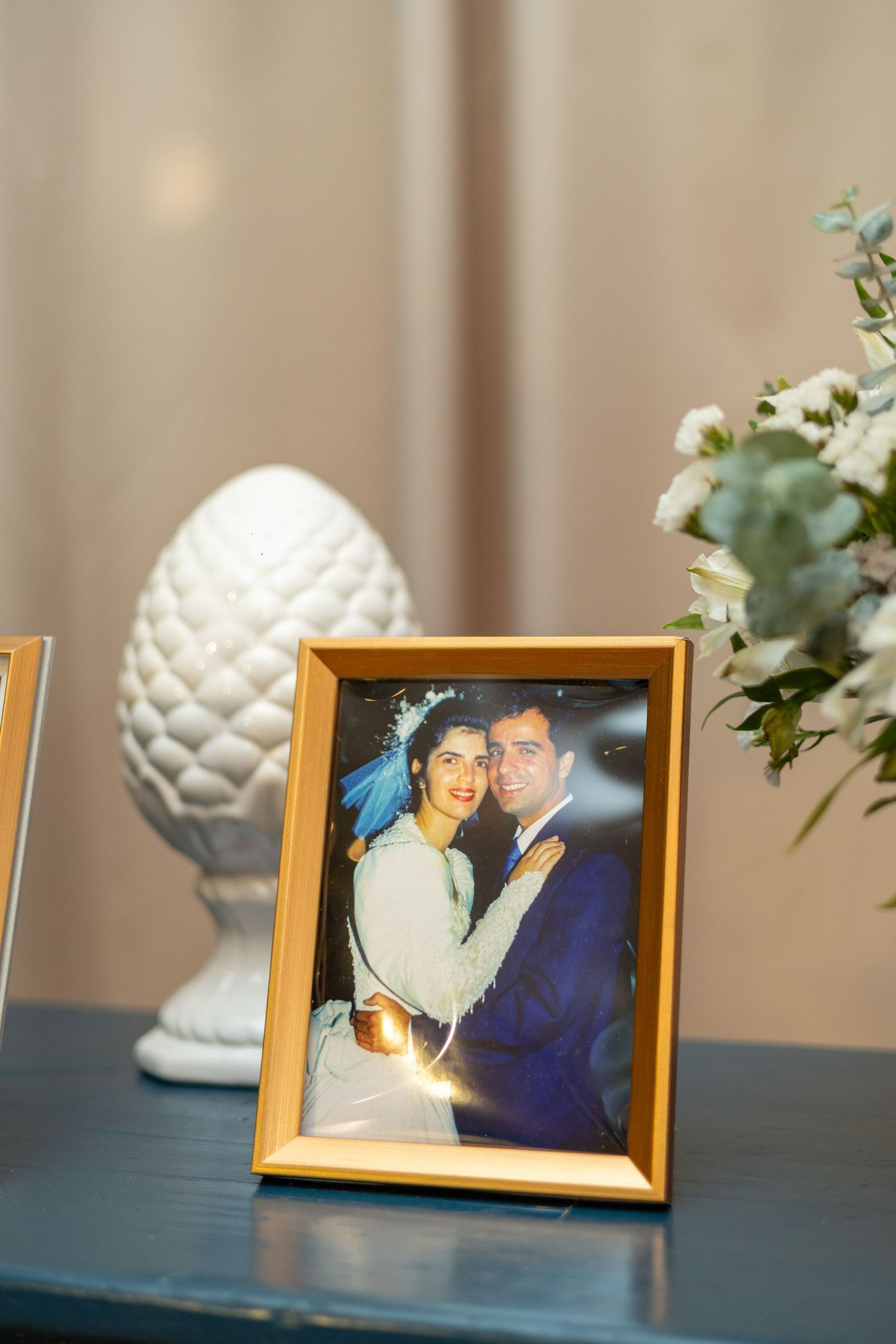 Gold framed wedding photo of a couple displayed on a table with white floral arrangement and decorative pinecone figurine.