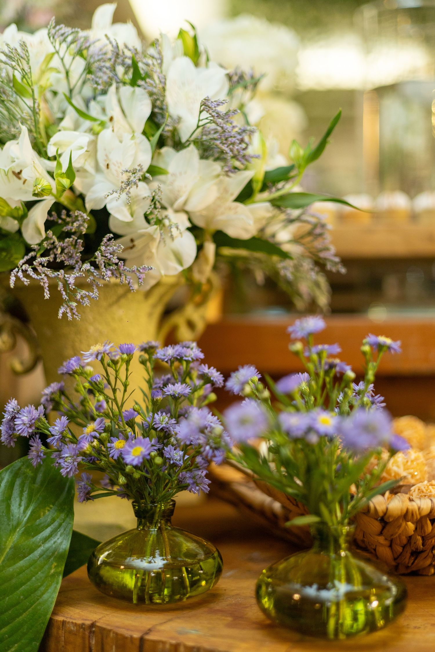 Purple aster flowers in small green glass vases beside a large white floral arrangement on a wooden table.