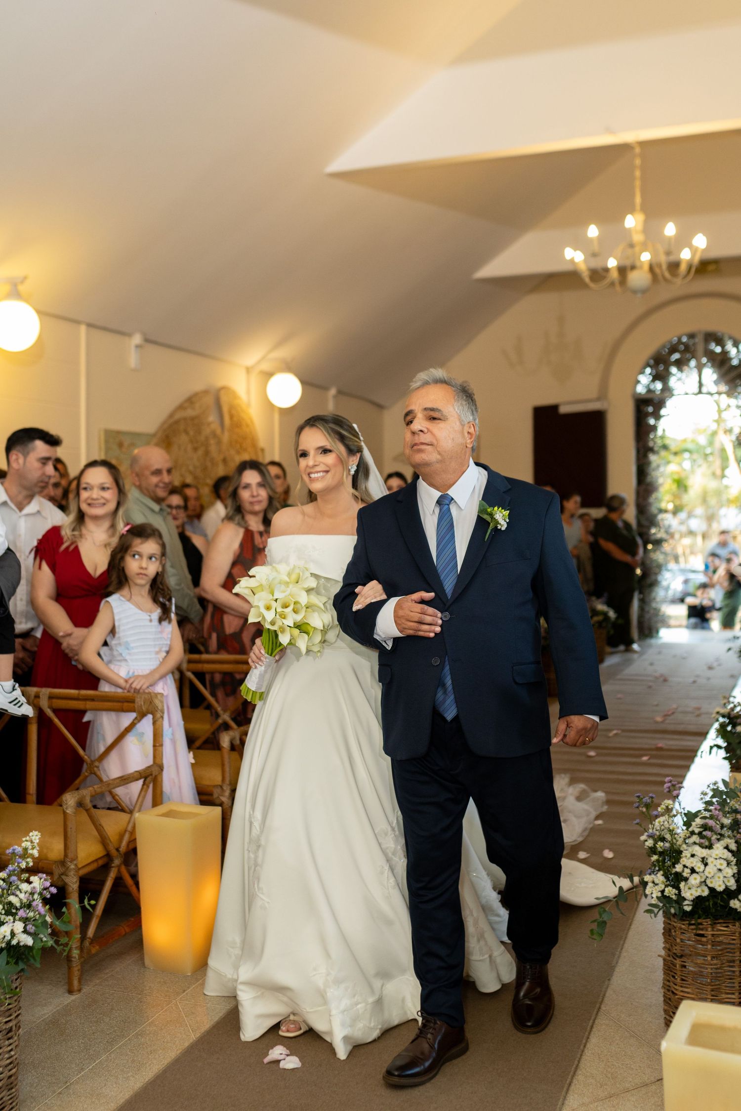 Bride in white gown walks down church aisle with escort in navy suit, guests seated alongside floral arrangements.