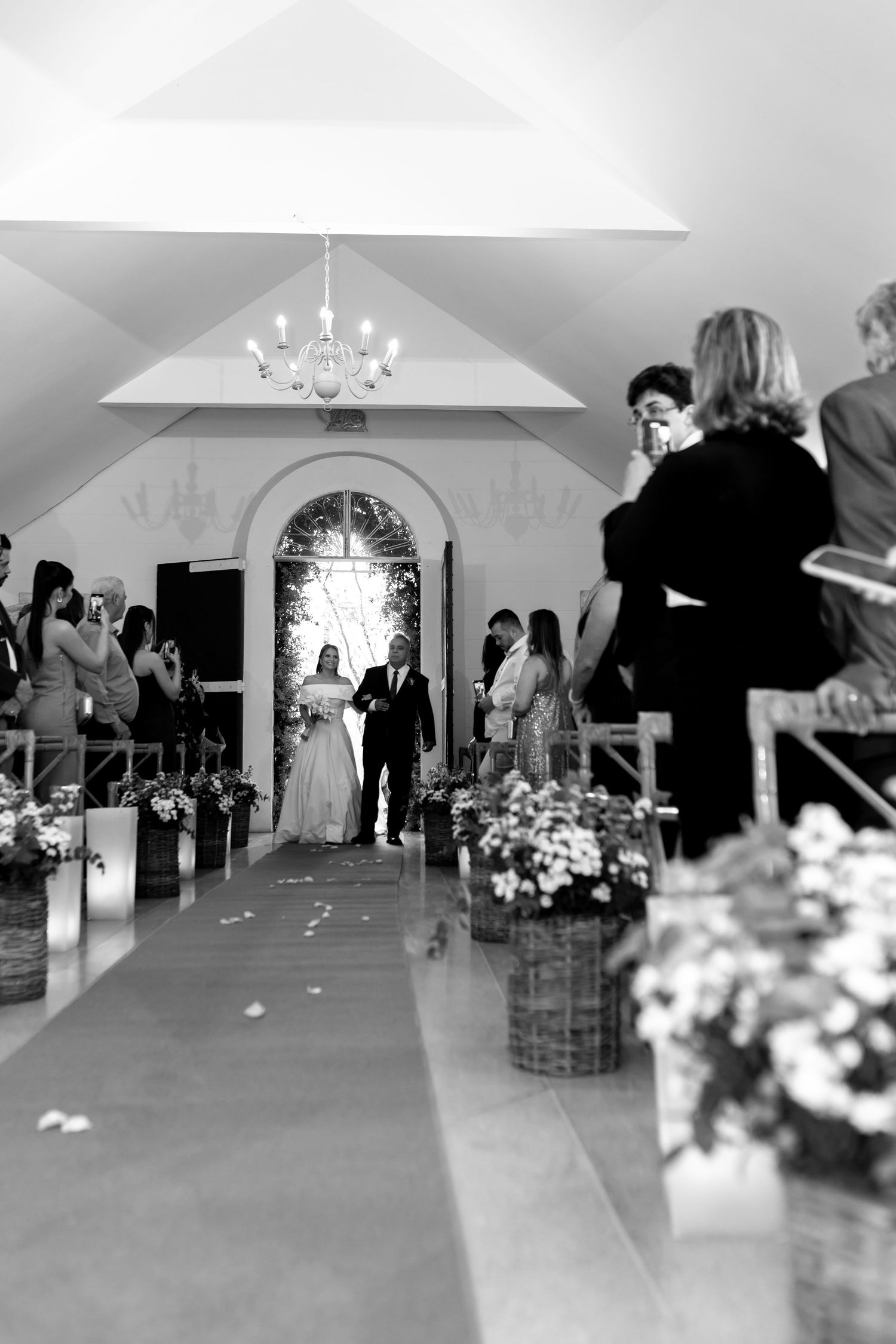 Black and white wedding ceremony photo showing bride walking down aisle with chandelier and floral arrangements.