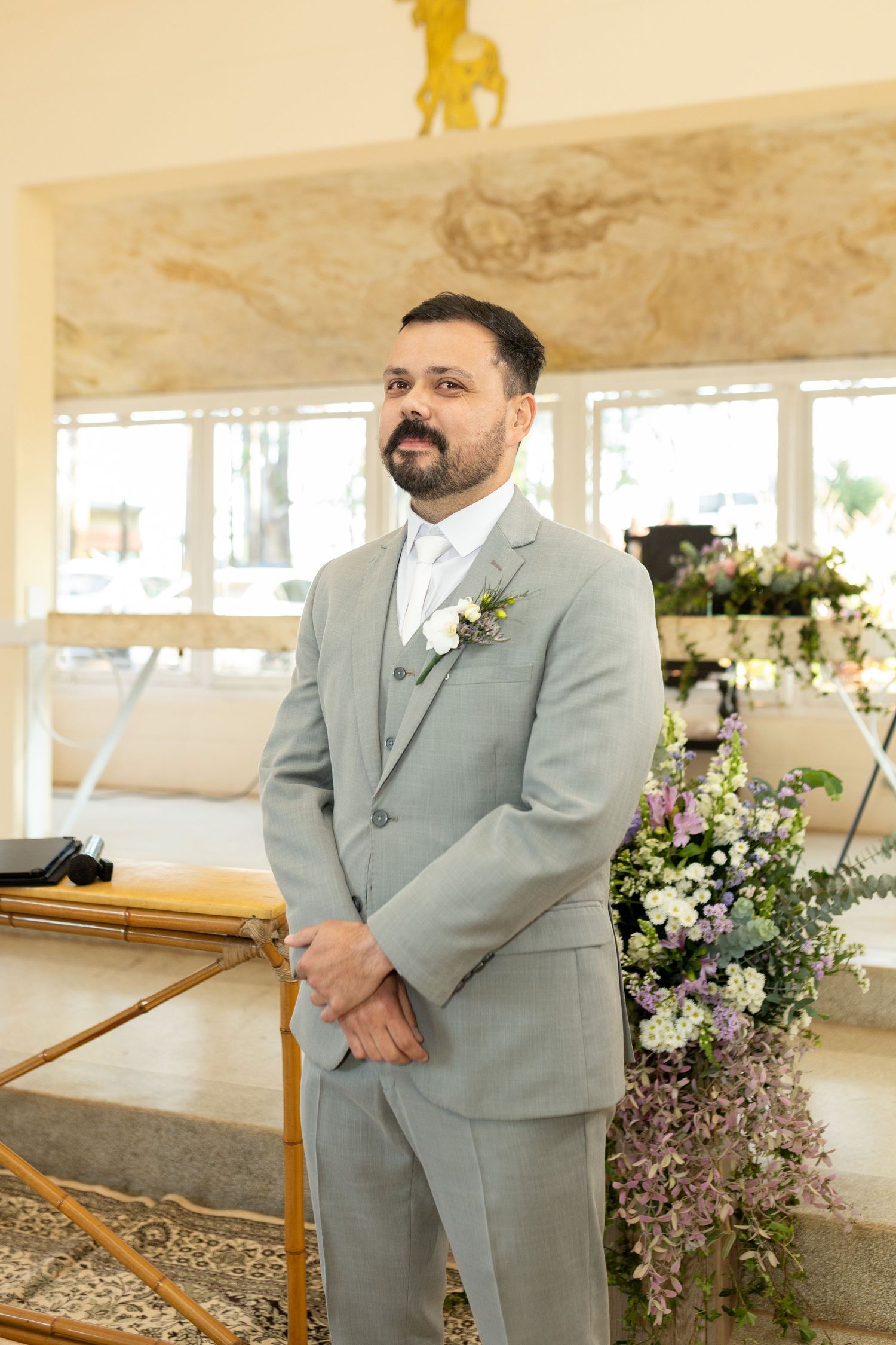 Groom in gray suit with boutonniere stands at wedding altar surrounded by floral arrangements.