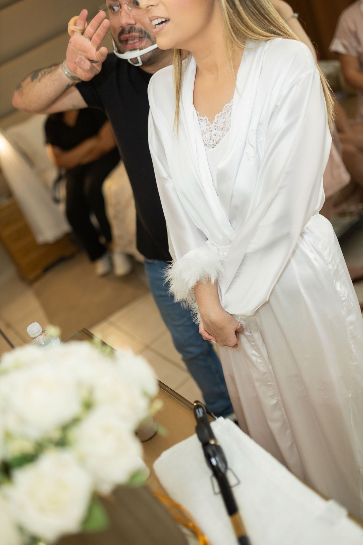 Bride in white satin robe with feather trim stands near white flowers while man in black shirt gestures nearby.