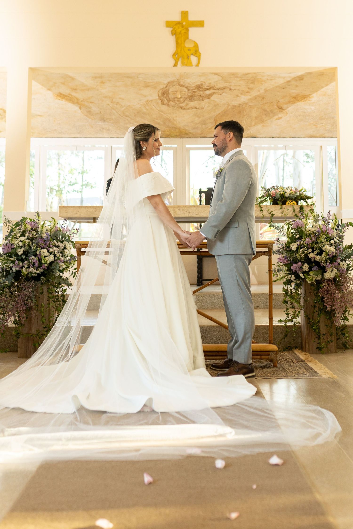Bride and groom hold hands at church altar during wedding ceremony, with floral arrangements and gold cross.