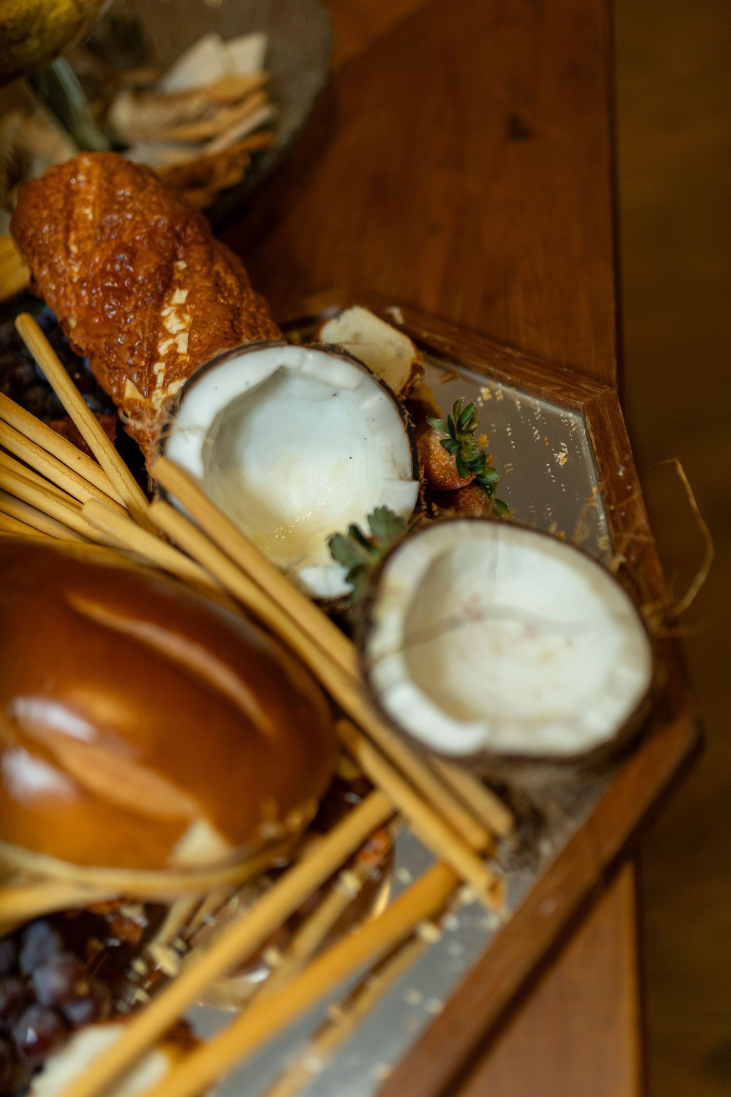 Rustic wooden tray with halved coconut, breadsticks, golden bread rolls, and assorted snacks on a wooden table.