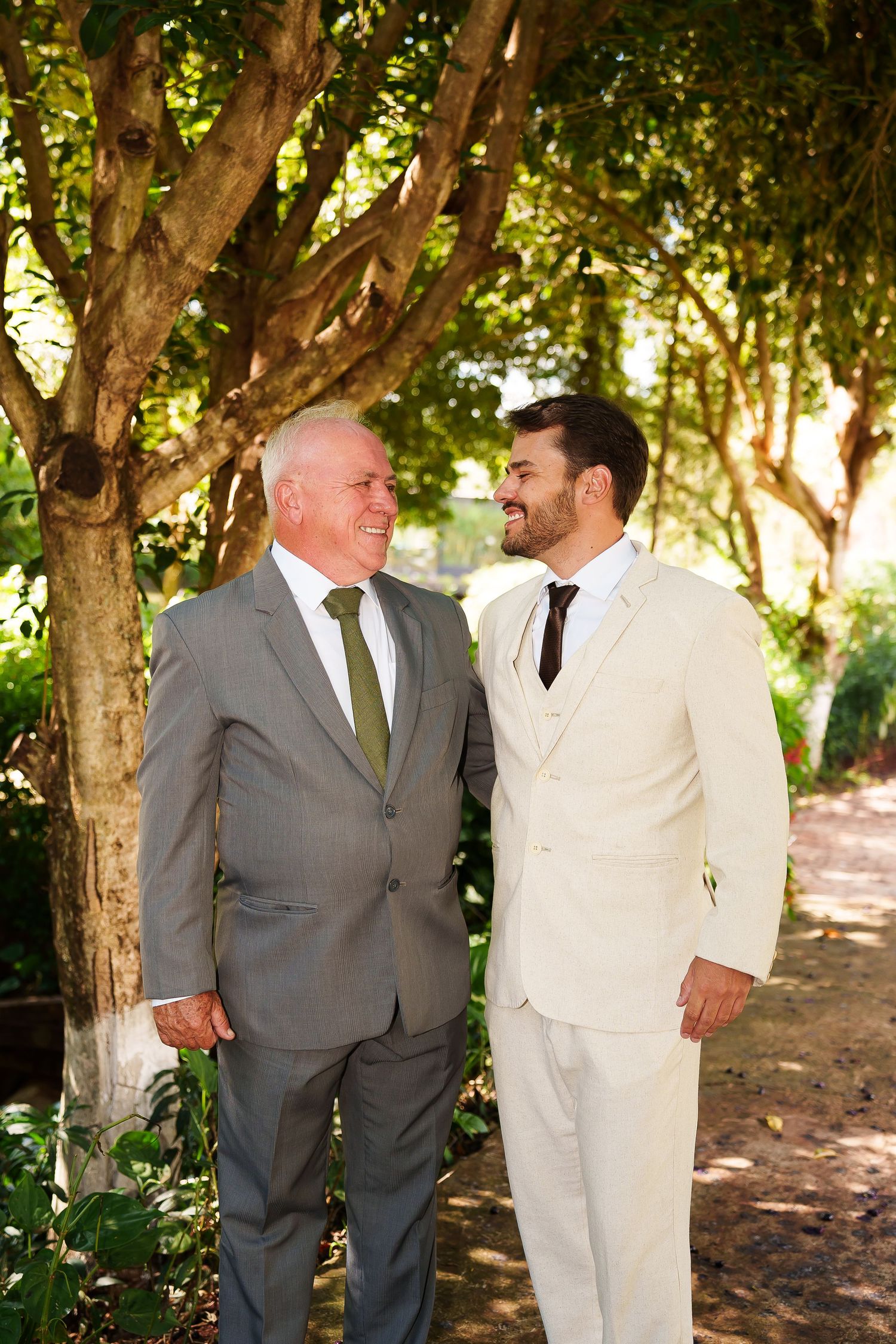 Two men in suits smiling at each other outdoors among green trees at a wedding celebration.