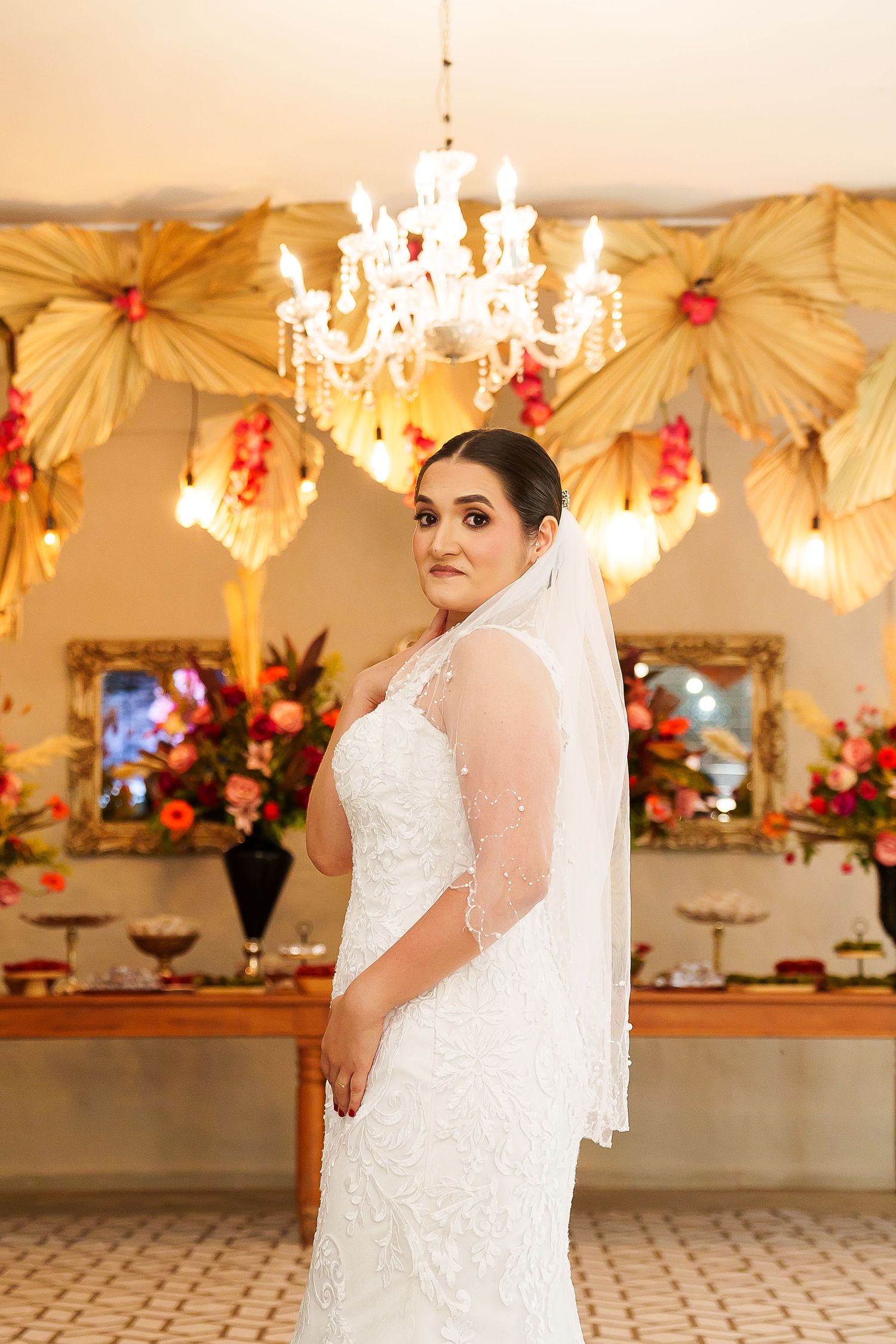 Bride in white lace gown and veil poses in elegant decorated venue with chandelier and floral arrangements.