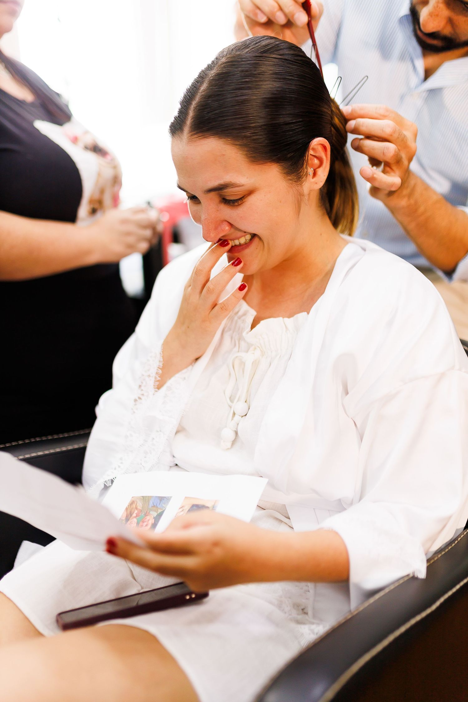 A smiling woman in a white robe reads a card while getting her hair styled at a salon.