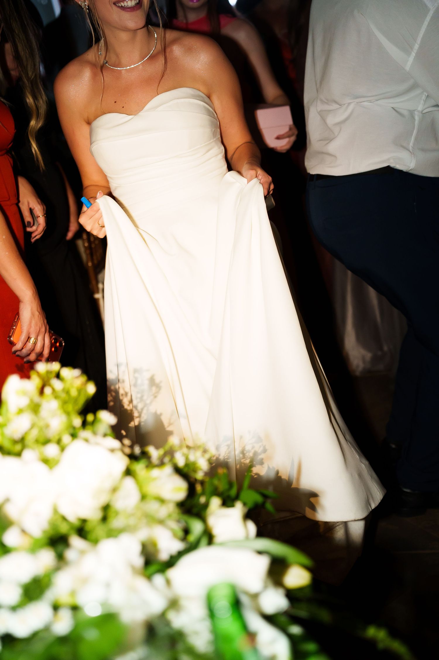 Bride in strapless white wedding gown dancing joyfully at reception with floral arrangements in foreground.