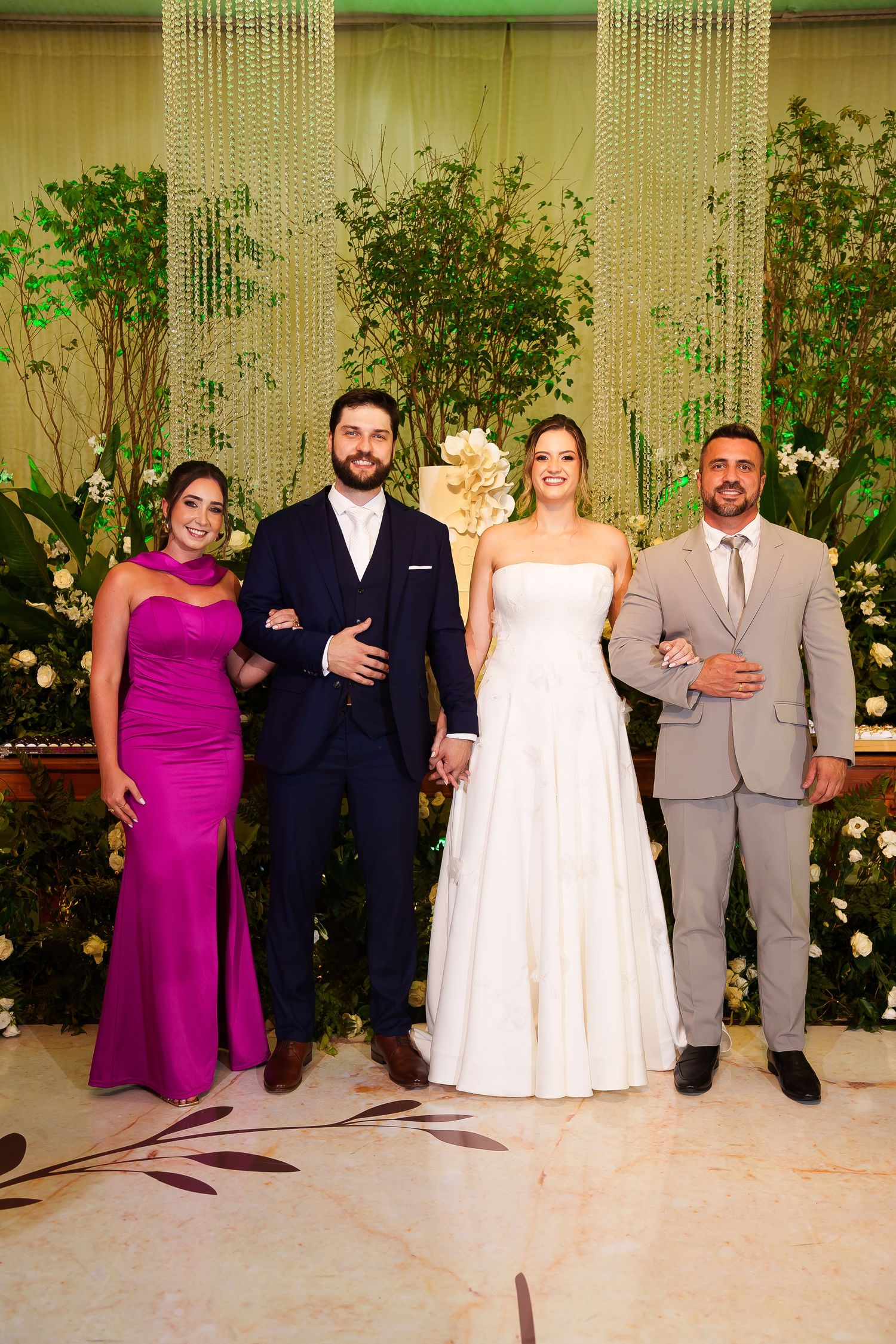 Wedding party poses elegantly in front of lush floral backdrop, bride in white gown with groom and guests.