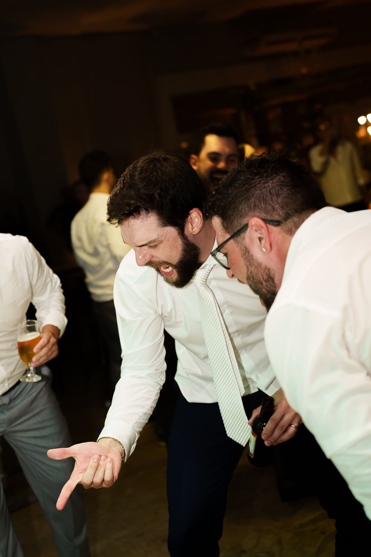 Men in white dress shirts and ties laughing and dancing together at a wedding reception celebration.