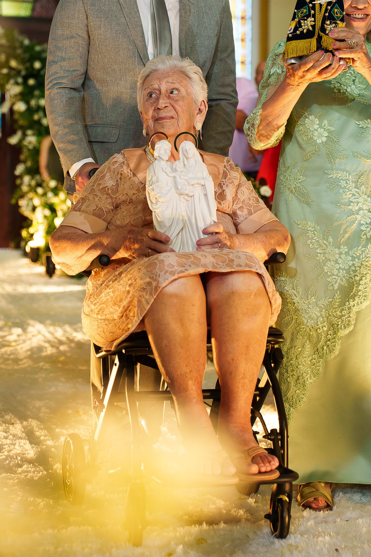 Elderly woman in wheelchair holding white angel statue at formal event, wearing beige lace dress, surrounded by guests.