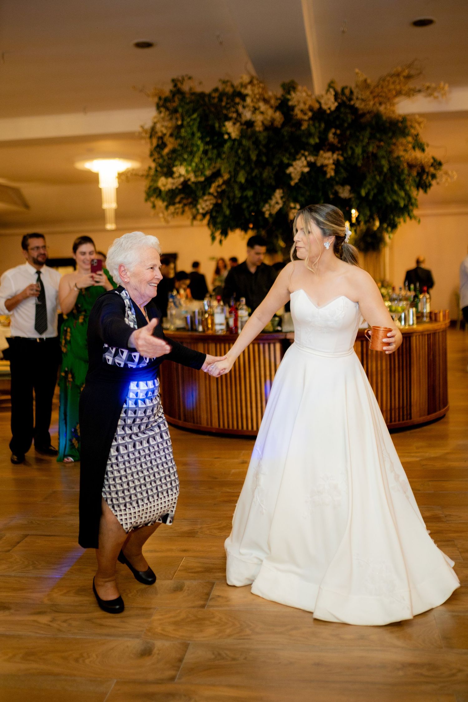 Bride in white gown dancing joyfully with elderly woman in patterned dress at elegant wedding reception.