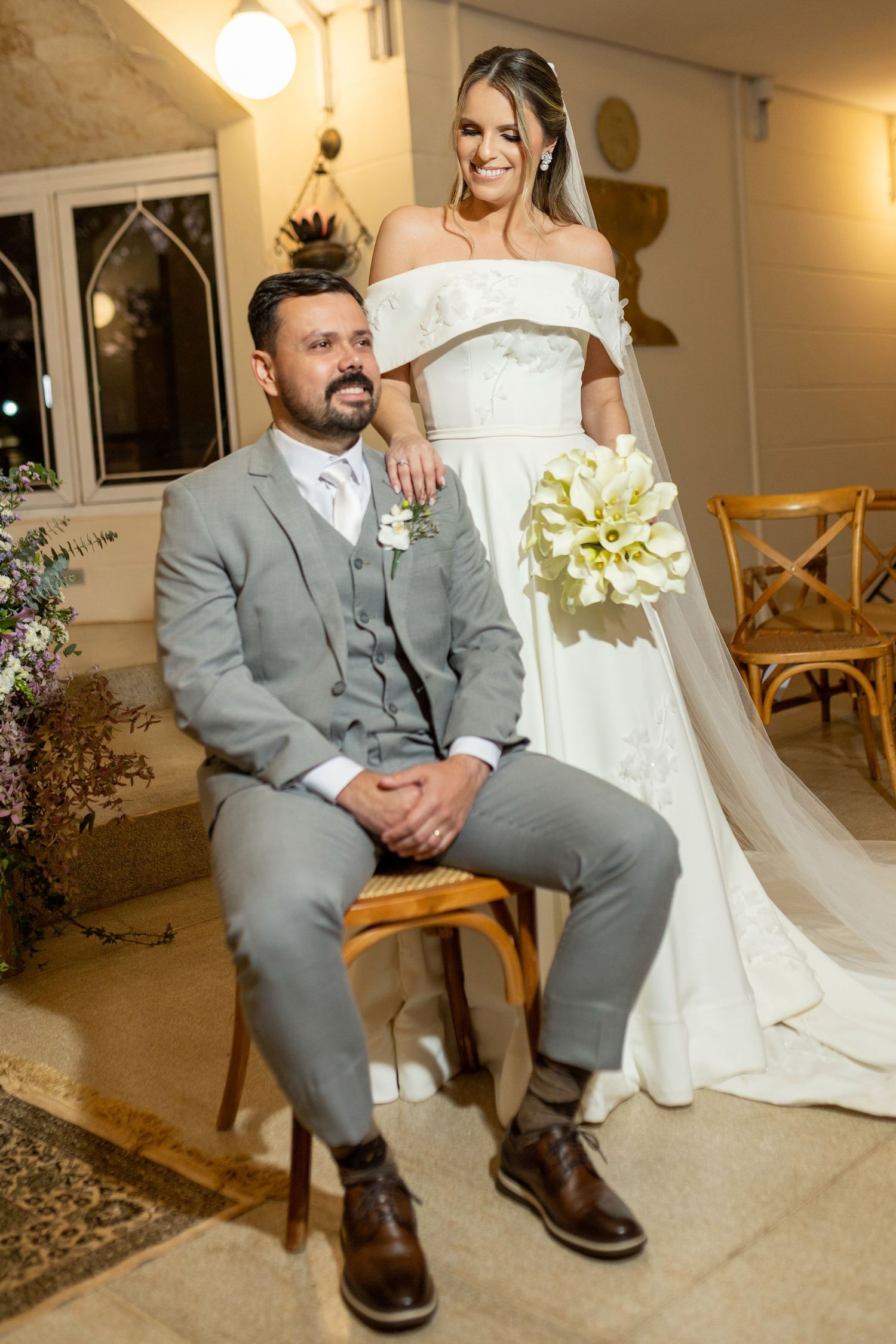 Bride in white off-shoulder gown holding calla lilies stands beside seated groom in gray suit at elegant wedding venue.