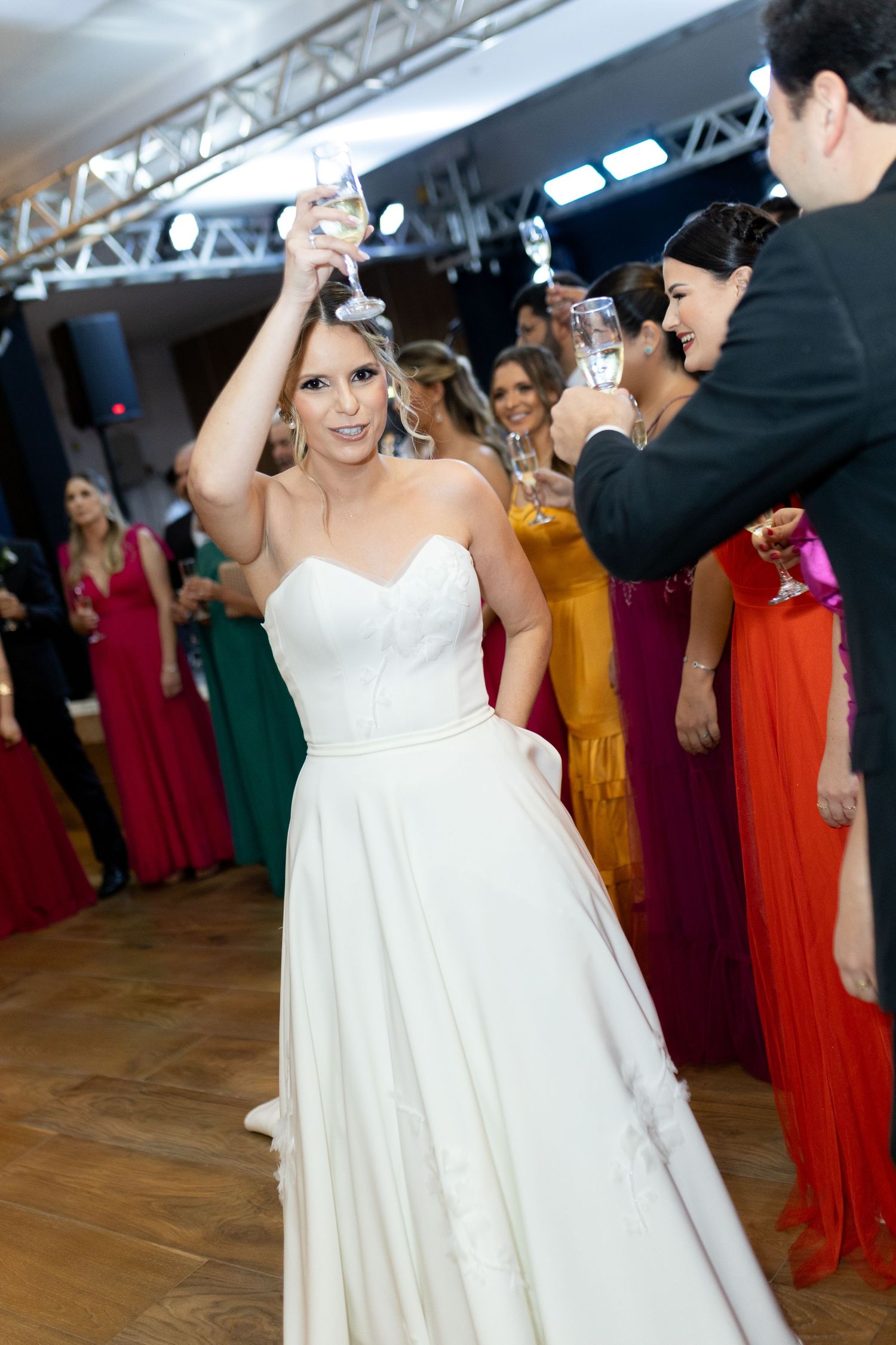 Bride in white strapless gown raises champagne glass during wedding reception toast with colorful guests.