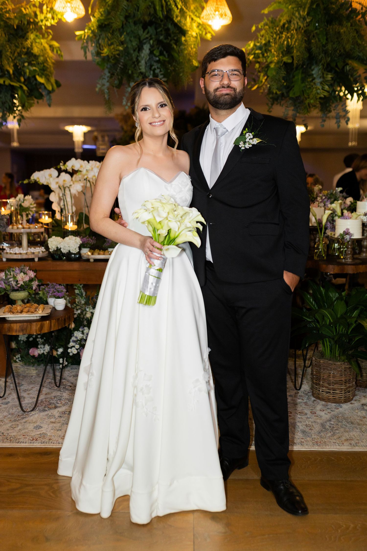 Bride in white gown holding calla lily bouquet stands with groom in black suit at elegant floral wedding reception.