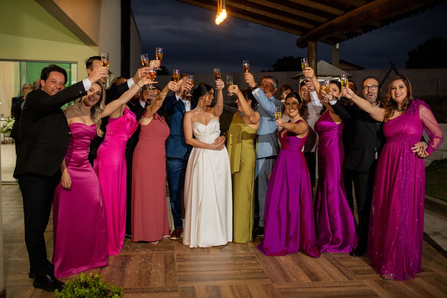 Wedding party celebrating outdoors at night, bride in white gown surrounded by guests in vibrant pink and colorful formal attire.