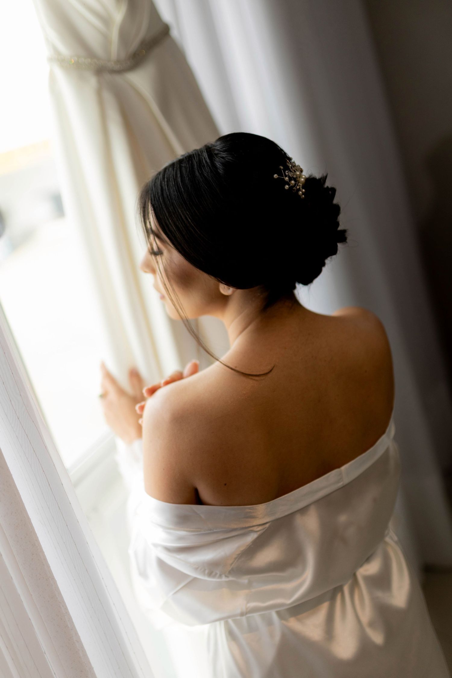 Bride with dark hair updo and floral accessory in white off-shoulder gown gazing out a sunlit window.