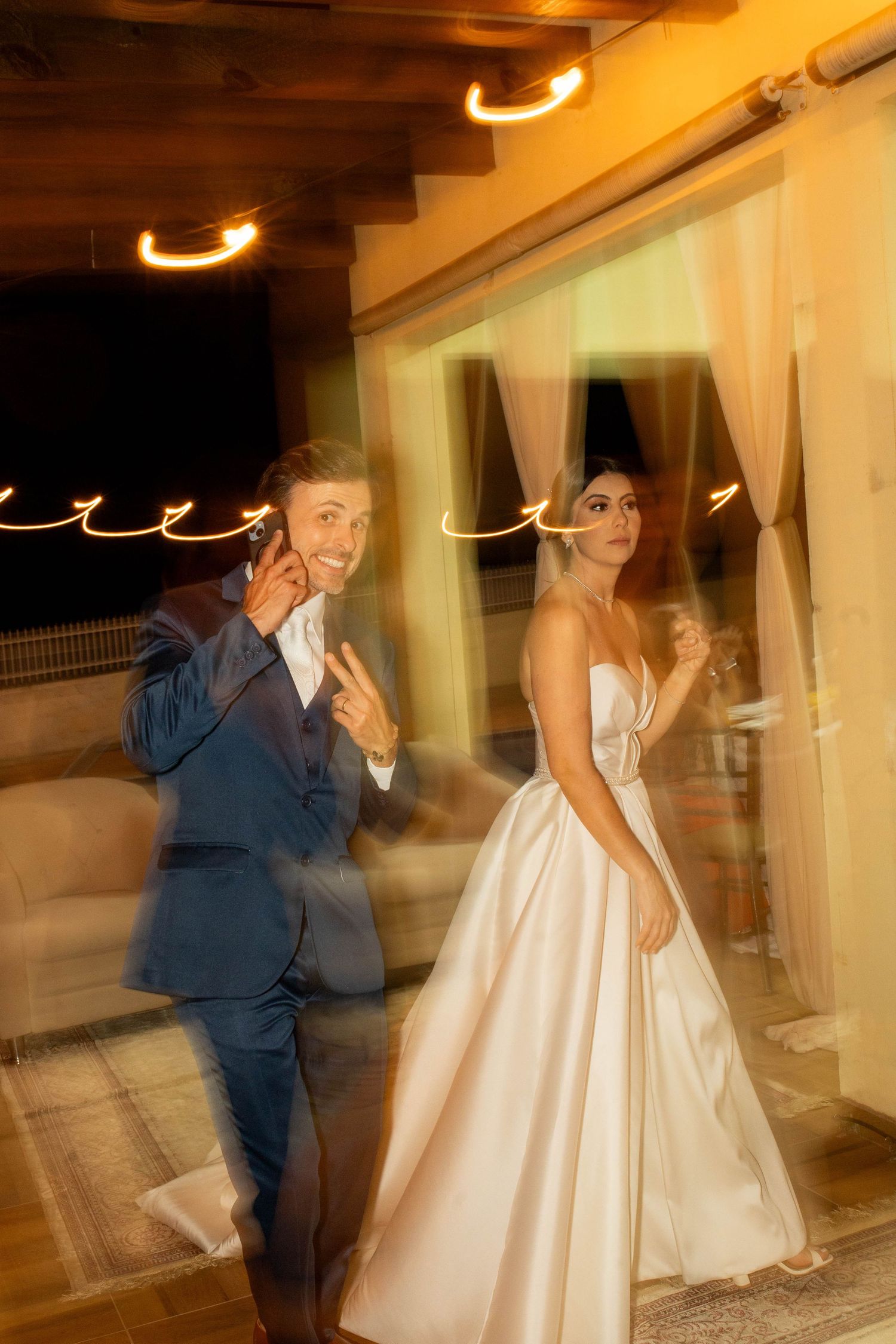 Bride in white gown and groom in blue suit celebrate at wedding reception with warm ambient lighting.