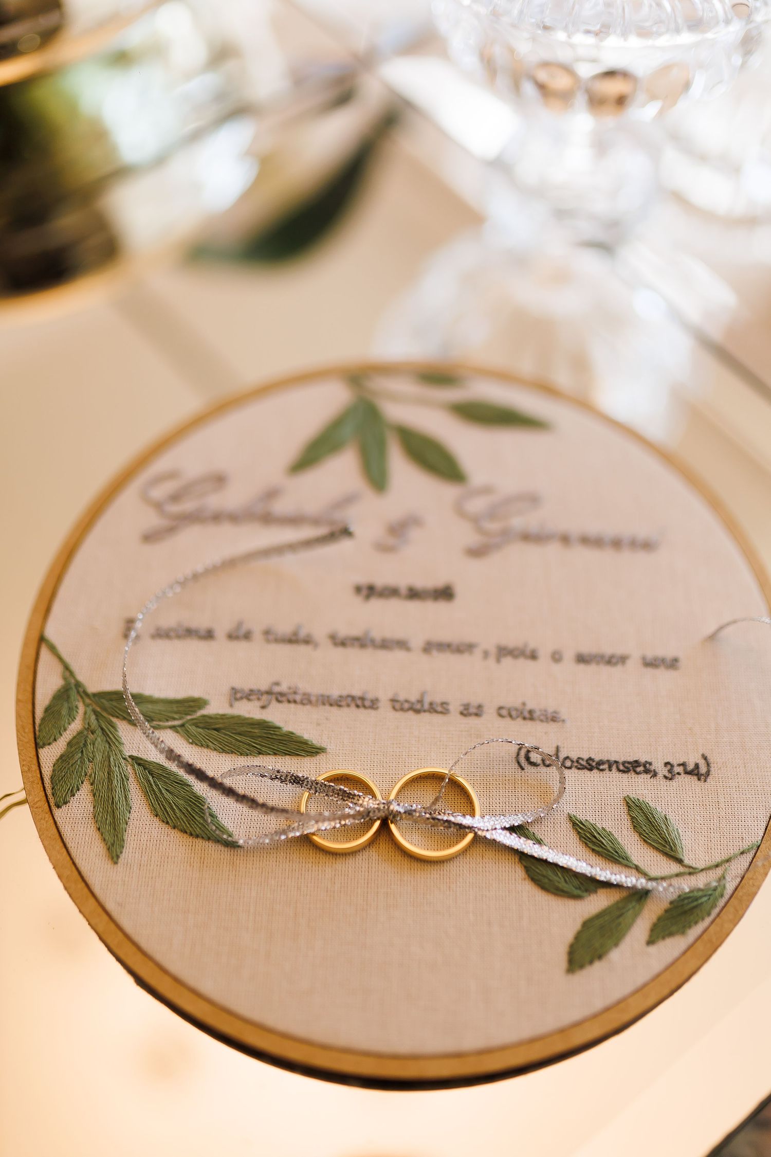 Two gold wedding rings on an embroidered hoop with floral design and Colossians 3:14 scripture verse.