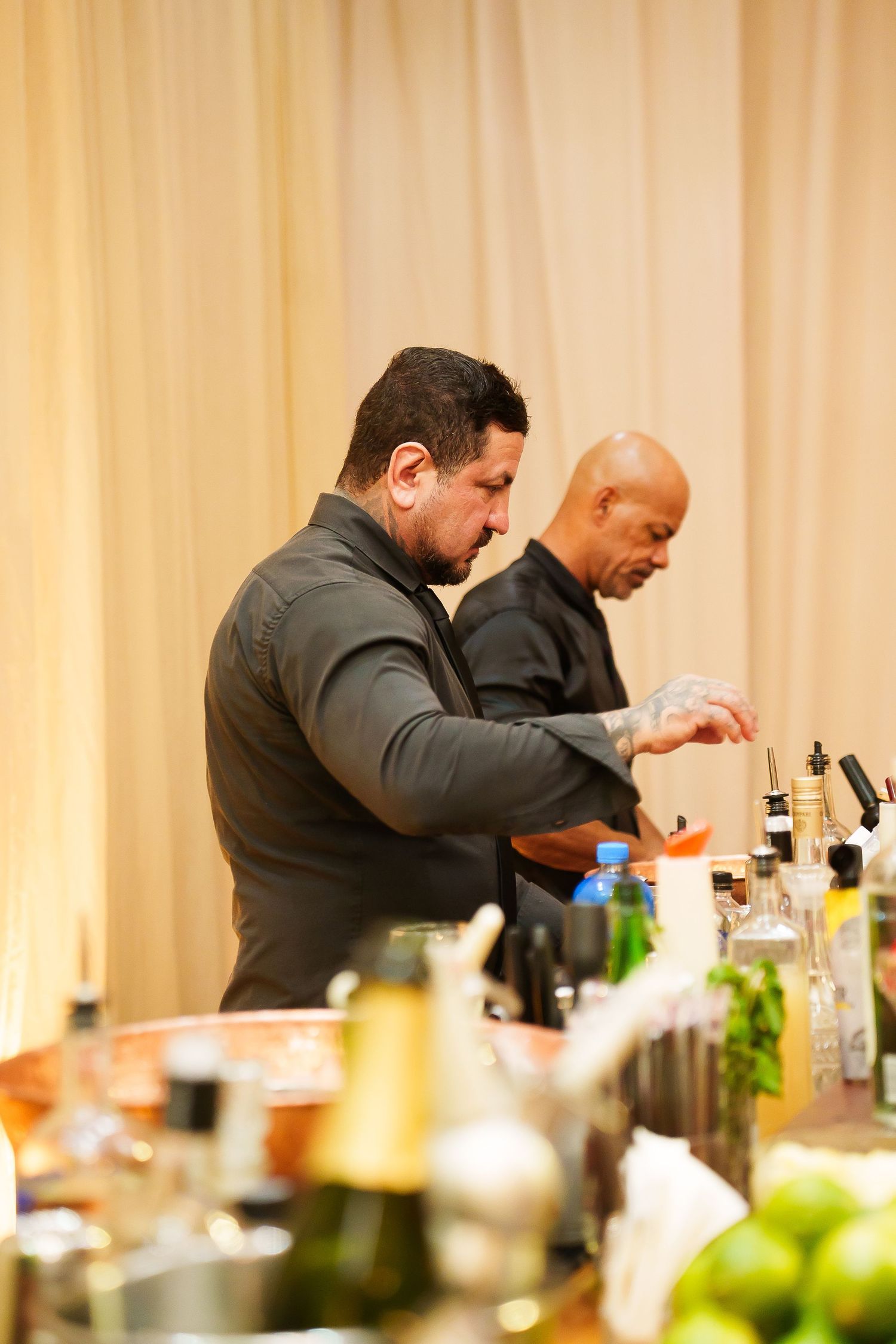 Two bartenders in dark shirts preparing cocktails at a bar with bottles and garnishes in the foreground.