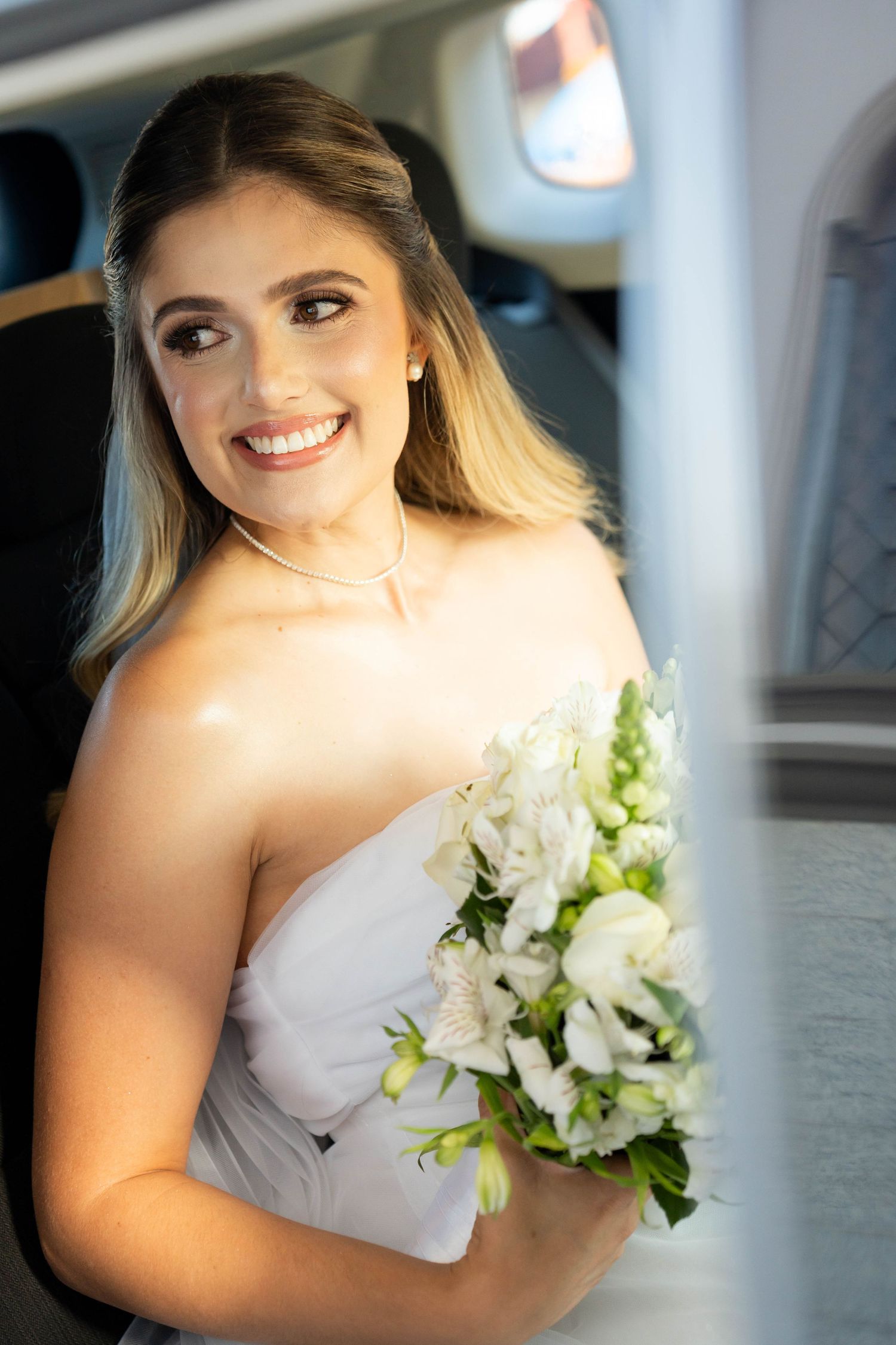 Smiling bride in strapless white dress holding bouquet of white flowers while sitting in a car on her wedding day.