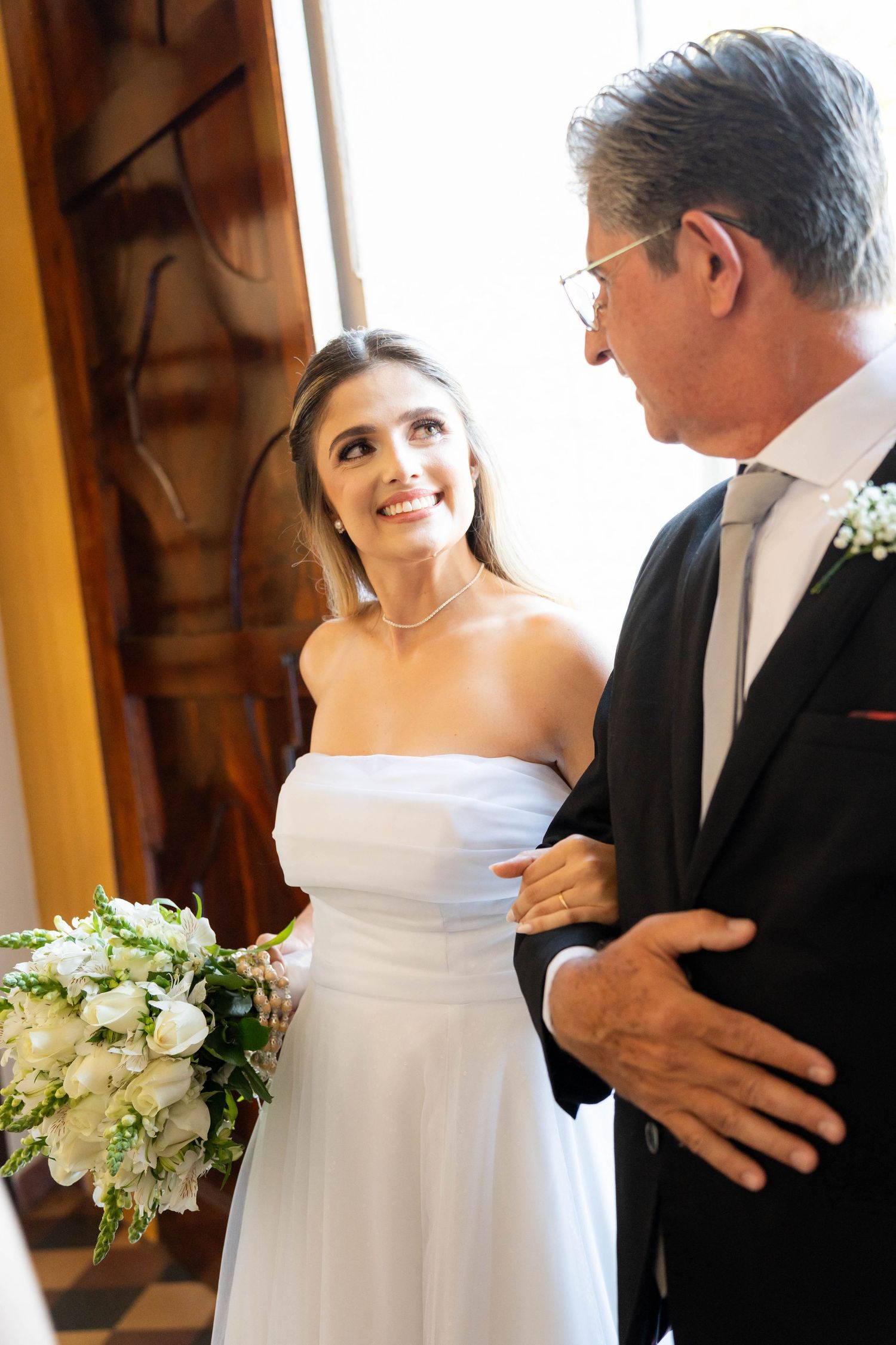 Smiling bride in white dress holding bouquet, standing with older man in black suit at wedding ceremony.