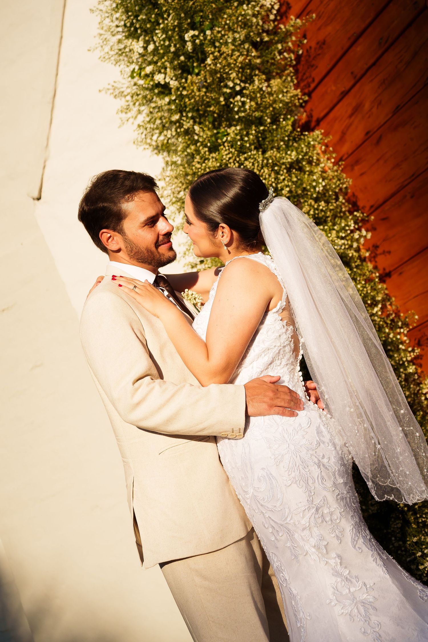 A bride and groom share a romantic embrace outdoors, with the bride in a lace gown and veil, groom in a beige suit.