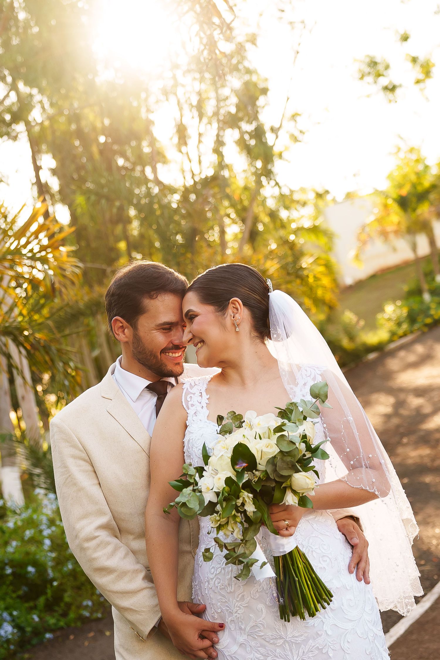 Bride and groom share a romantic moment outdoors, with bride holding white bouquet, bathed in golden sunlight.