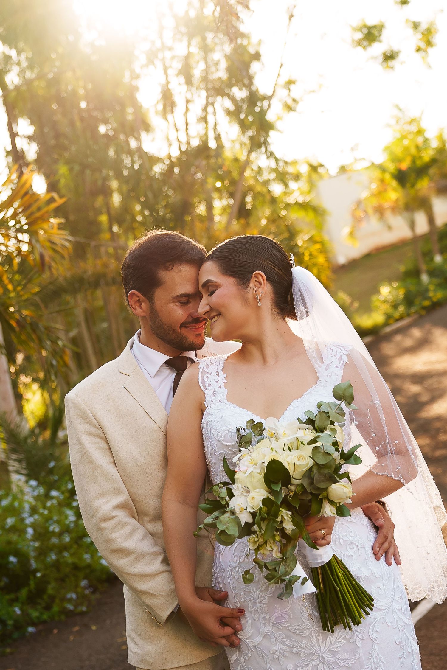 A bride and groom share a romantic forehead touch outdoors, she holds a white bouquet, bathed in golden sunlight.