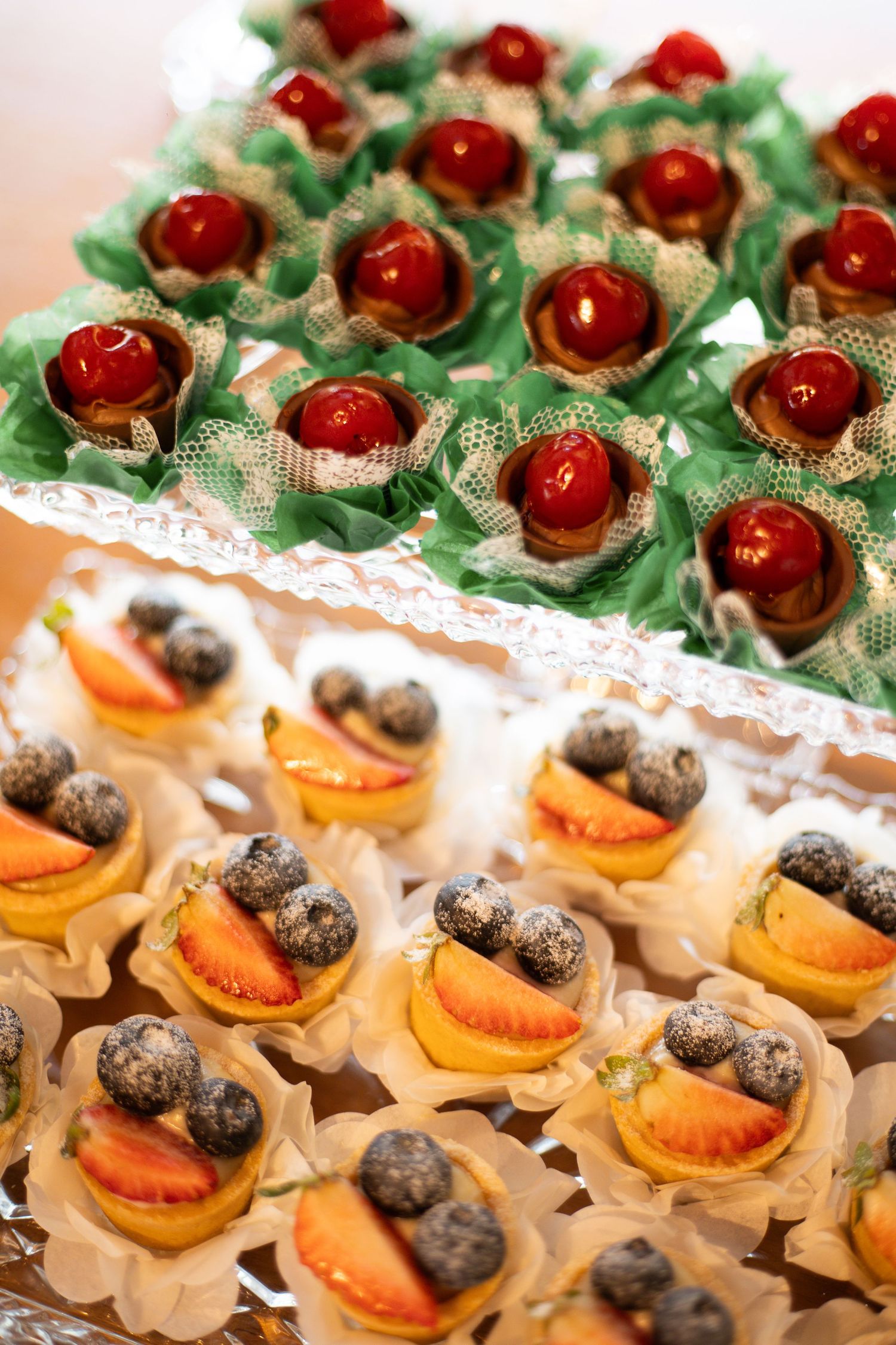Elegant dessert trays with fruit tarts topped with strawberries and blueberries, and cherry chocolate cups on green lace.