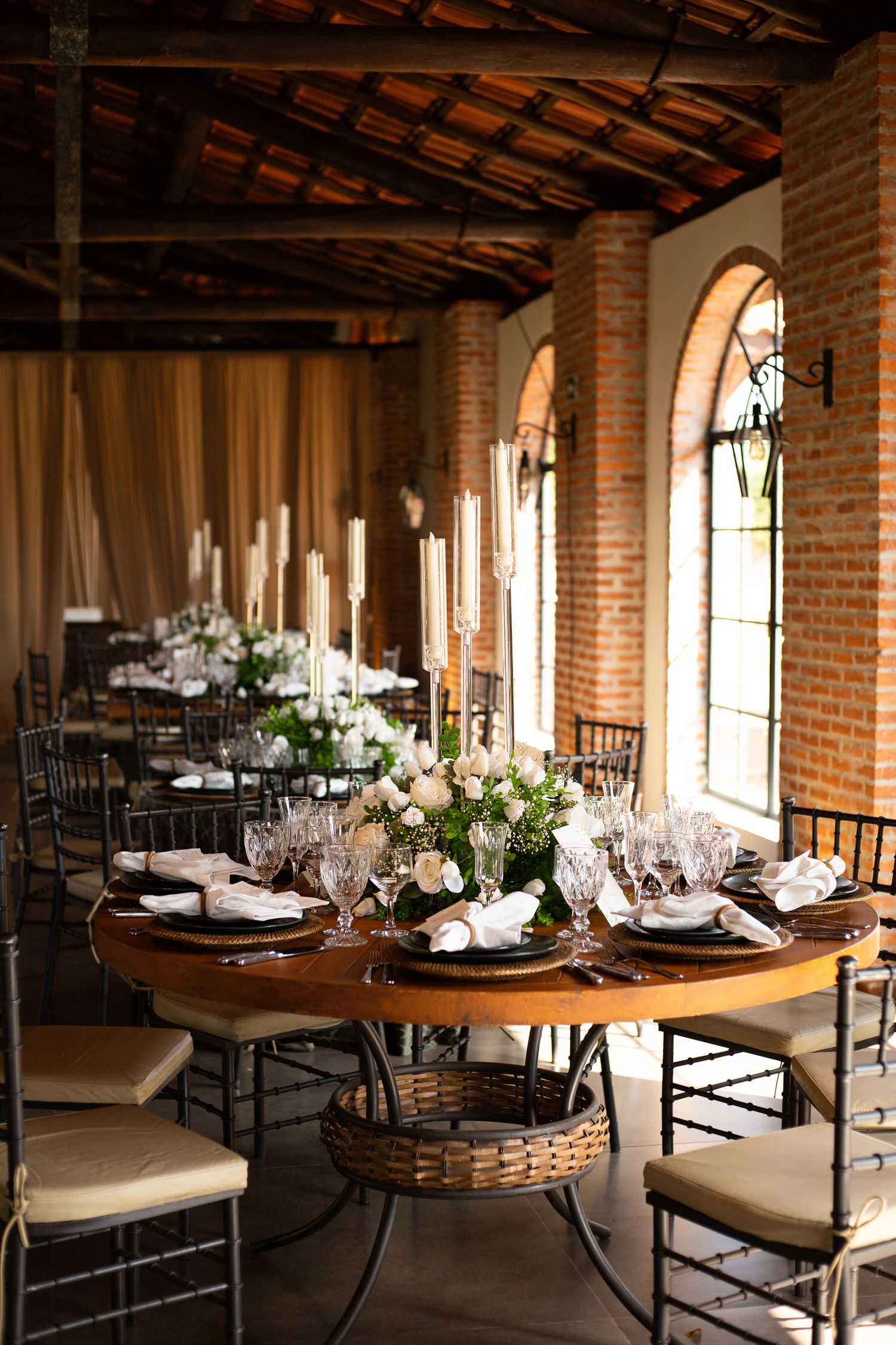 Elegant wedding reception hall with round wooden tables, crystal glasses, floral centerpieces, and arched windows in a rustic brick venue.