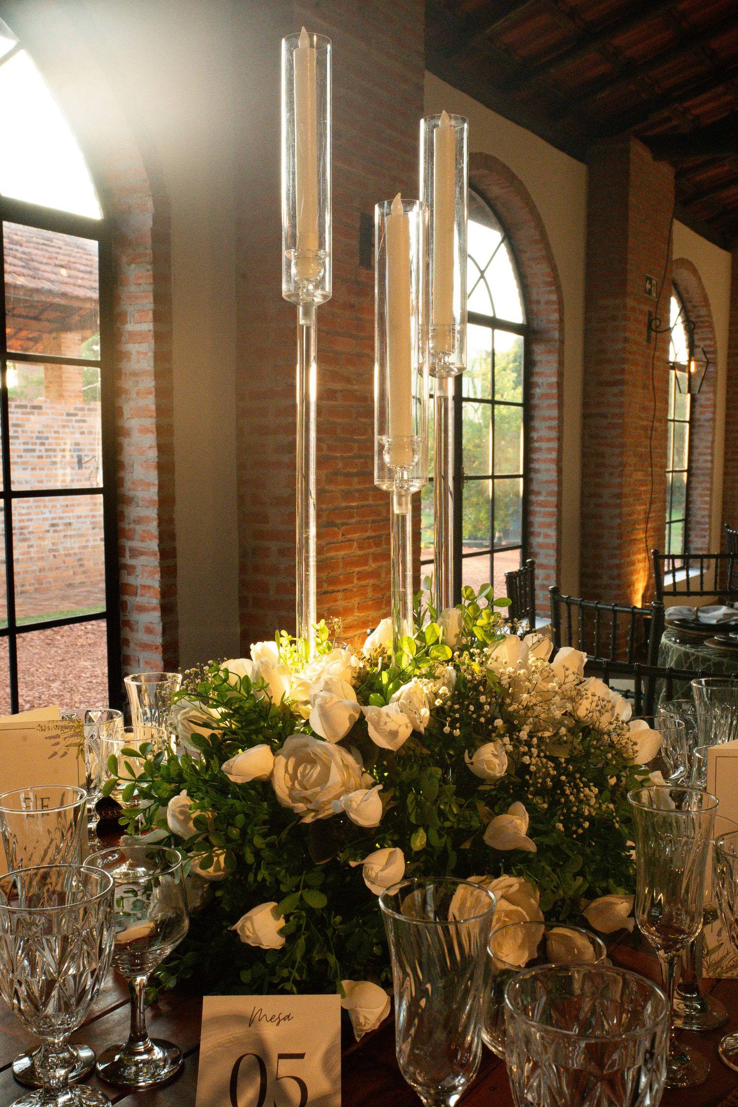 Elegant wedding centerpiece with white roses, greenery, and tall glass candelabra on a crystal-adorned table in a brick venue.