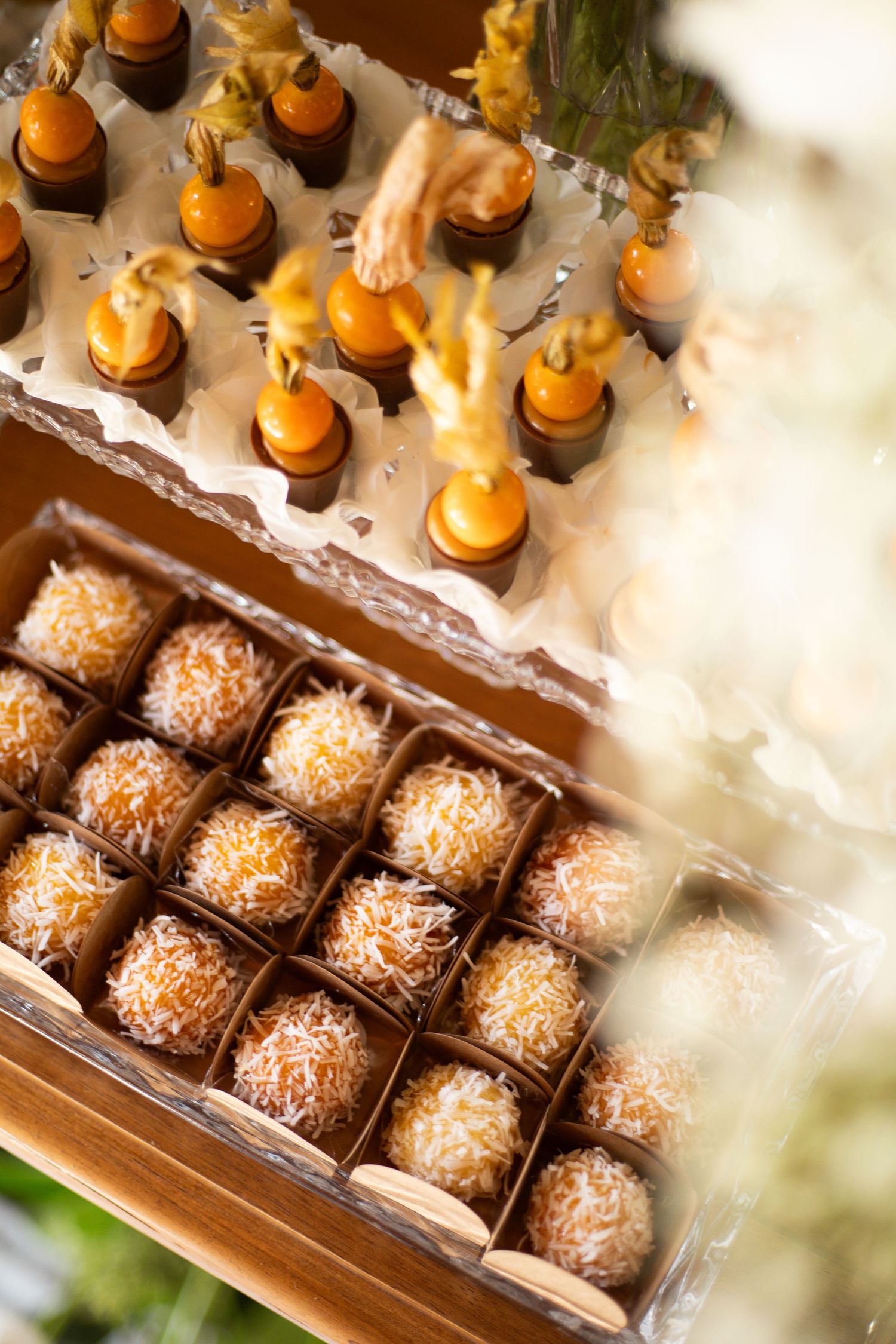 A wooden box display of coconut-dusted truffles and orange chocolate bonbons topped with edible gold flowers at a catered event.