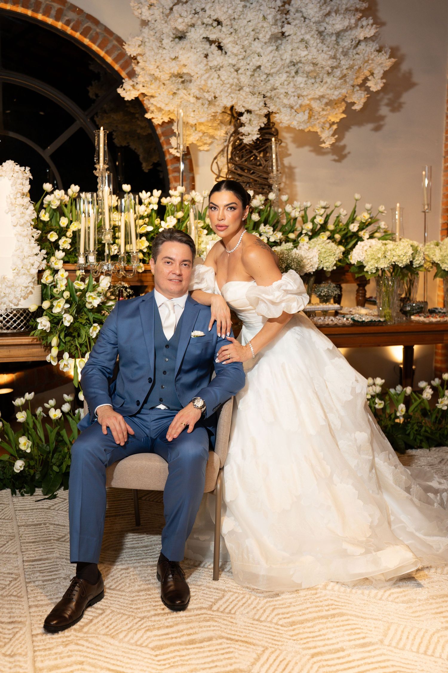 Elegant wedding couple posing at floral reception, groom in blue suit, bride in white off-shoulder ballgown with white flower backdrop.