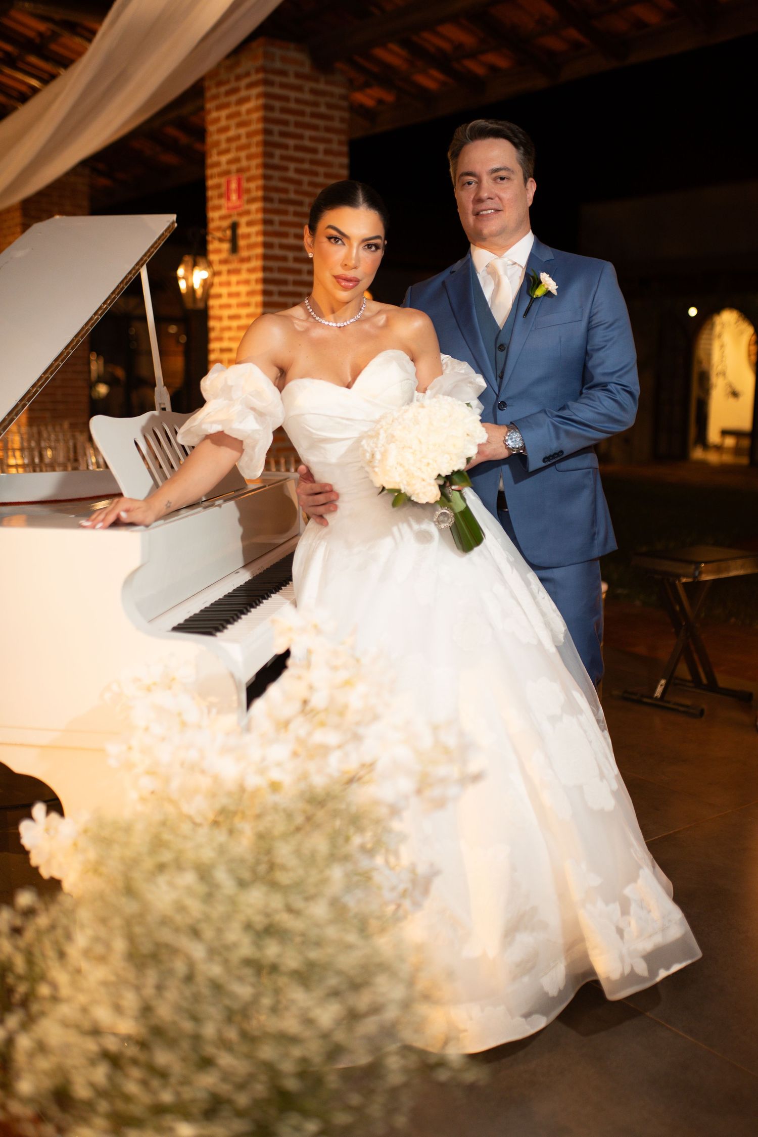 Bride in white ball gown poses with groom in blue suit beside a white grand piano at elegant wedding venue.